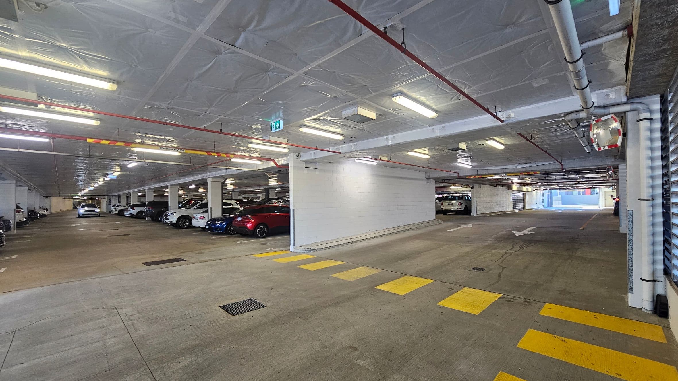 An underground car park with a concrete floor and a low ceiling lined with fluorescent lights and pipes. On the left, several parked cars are visible, including a white and a red vehicle. The right side features a white wall and more parked cars. Yellow pedestrian crossing lines are painted on the floor. In the background, the car park extends further with additional vehicles and an exit sign.