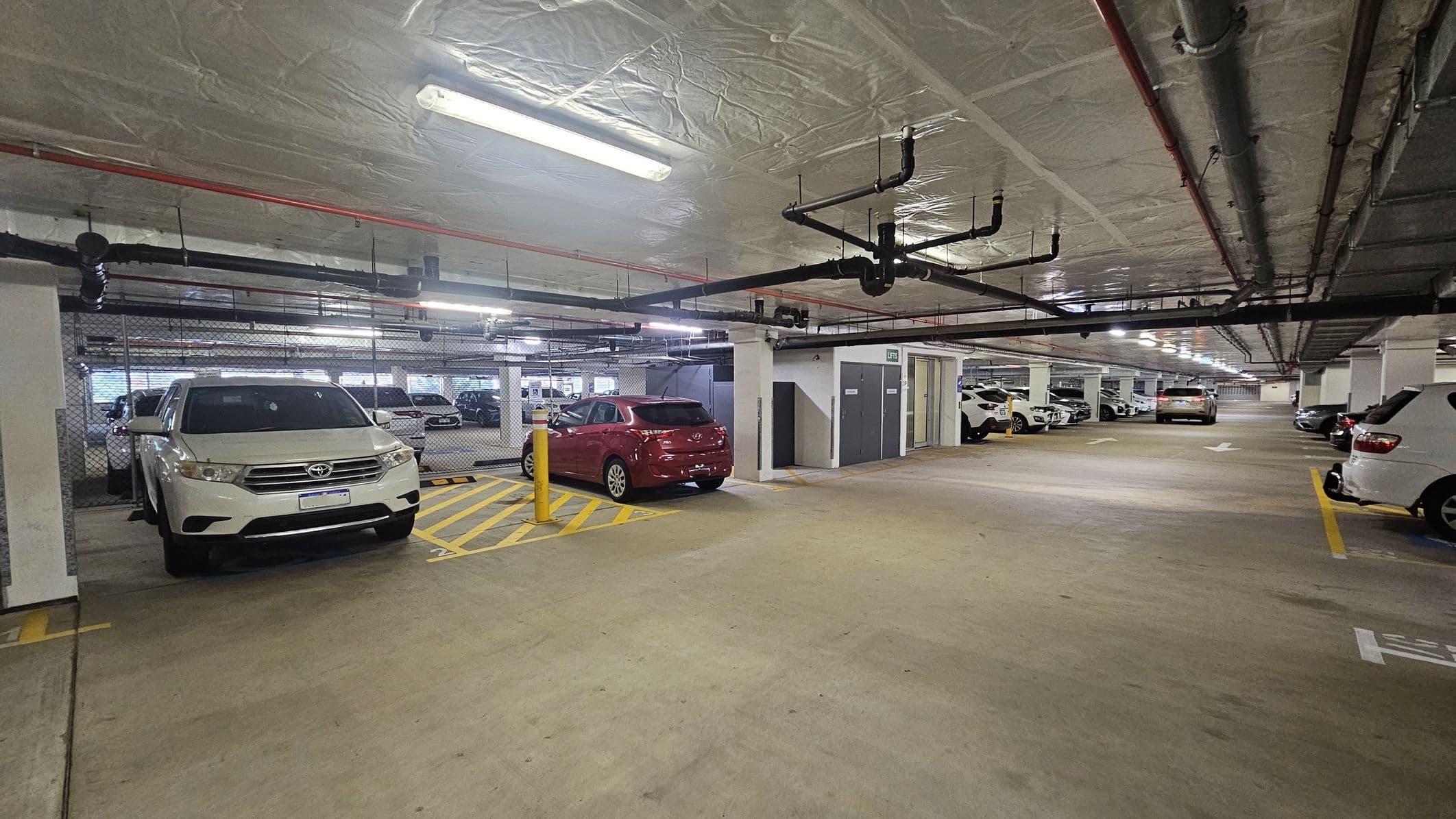 Underground car park with a concrete floor and a low ceiling lined with pipes and fluorescent lights. On the left, a white SUV is parked next to a red car, separated by yellow diagonal lines. The right side shows more parked cars along the wall. In the middle, the parking area extends into the distance with directional arrows on the floor. The walls are lined with metal fencing and there are storage lockers visible.