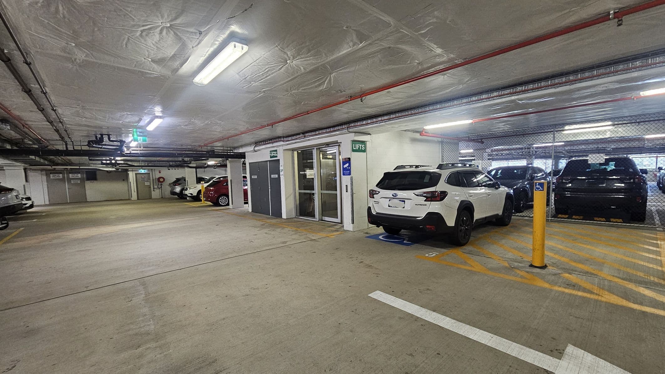 Underground car park with a concrete floor and ceiling. On the right, a white car is parked in a disabled parking space, marked by yellow lines and a sign. Adjacent is a glass door labelled "Lifts" with a green exit sign above. Several other cars are parked in the background, separated by a chain-link fence. Overhead, fluorescent lights and exposed pipes run along the ceiling. The area is well-lit and spacious.