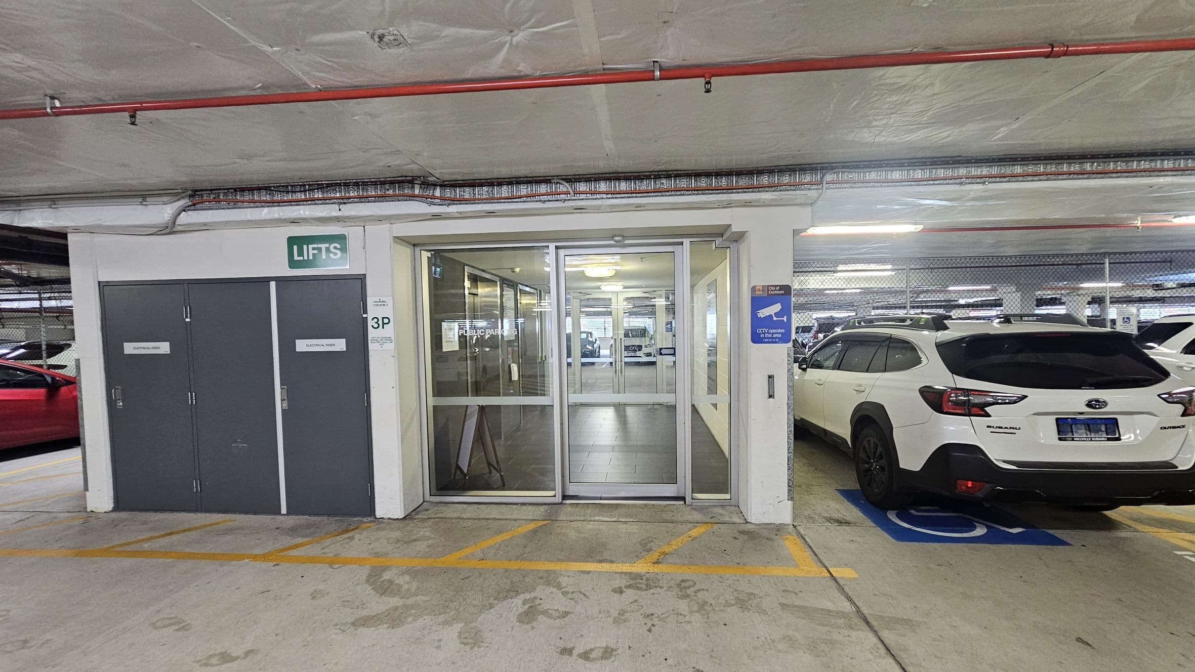A parking garage with a white car parked on the right in a designated space. In the centre, there are glass doors leading to a lift area, with a green sign above reading "LIFTS". On the left, there are two grey doors marked "ELECTRICAL RISER". The floor is concrete with yellow markings, and the ceiling is also concrete with visible pipes and wiring. A blue sign on the right indicates CCTV operation.