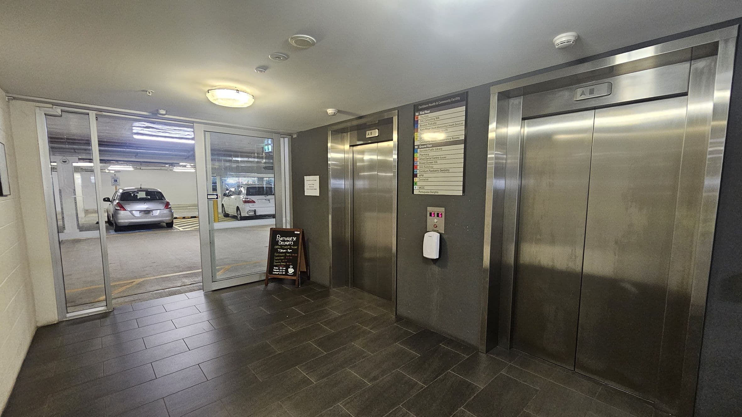 A hallway with two stainless steel lift doors on the right, each with a control panel and a directory sign above. In the middle, a small blackboard sign stands on the dark tiled floor. On the left, a glass wall reveals a car park with two parked cars. The ceiling is light-coloured with a circular light fixture.
