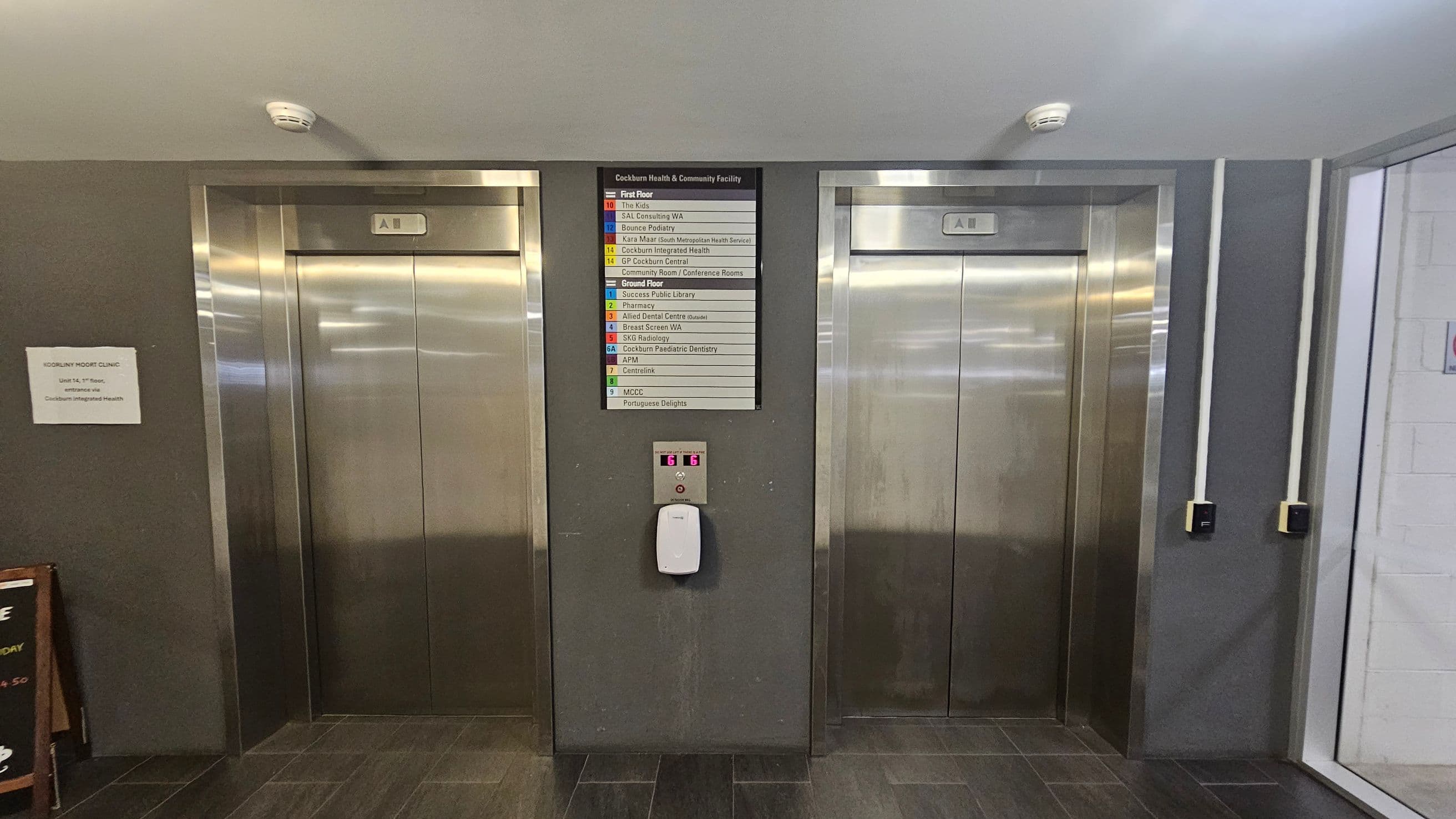 Two stainless steel lift doors are set into a grey wall. Between them, a directory sign lists various facilities, including a podiatry clinic and a library. Below the sign, a lift control panel with buttons and a hand sanitiser dispenser is mounted. On the left, a small sign indicates a podiatry clinic. The floor is tiled in dark grey, and the ceiling is light-coloured with smoke detectors. On the right, white pipes run vertically along the wall.