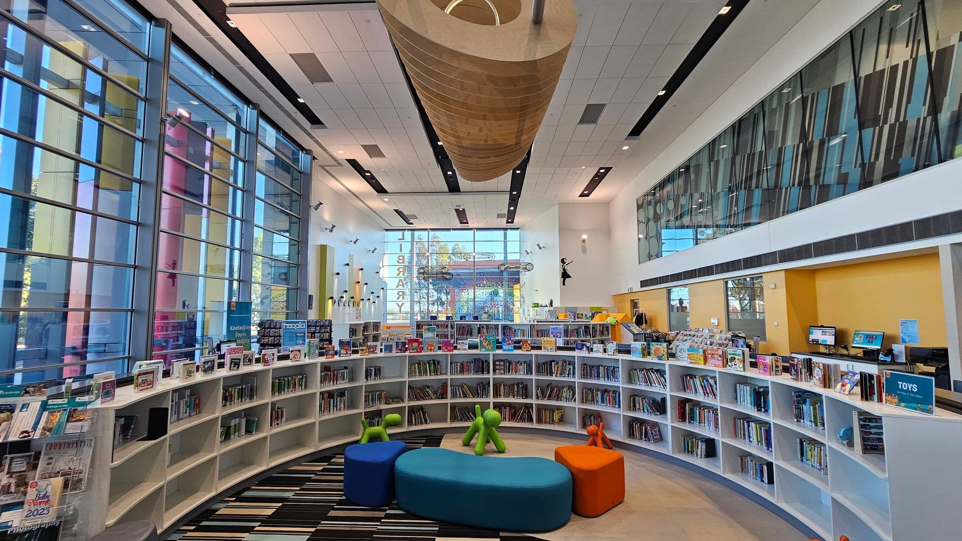 A modern library interior with large, colourful glass windows on the left, allowing natural light to flood in. In the centre, there are curved white bookshelves filled with books and displays. The foreground features colourful seating and playful, abstract figures. The ceiling is high with a large, wooden, cylindrical structure hanging from it. On the right, there are computers on a desk against a yellow wall. The floor is a mix of striped and plain patterns.