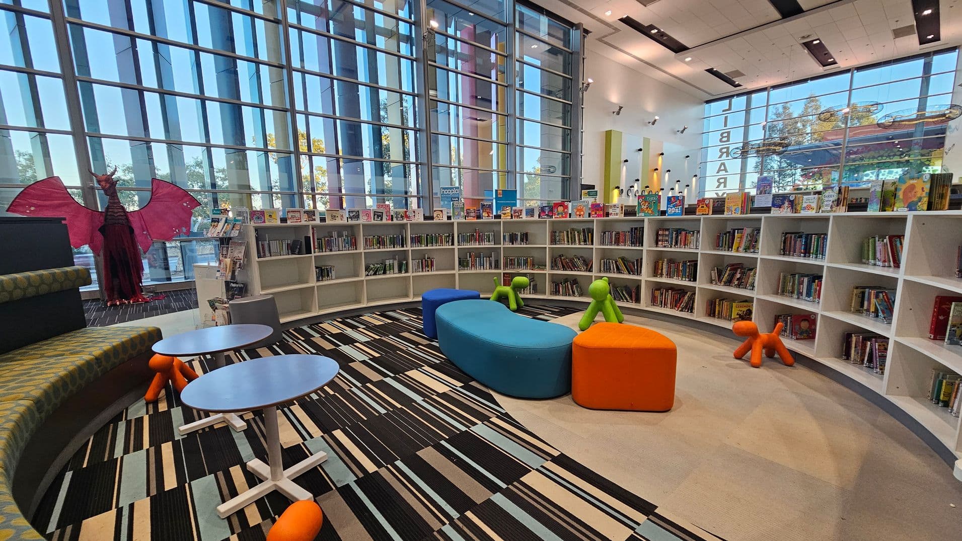A modern library interior with large floor-to-ceiling windows in the background, allowing natural light to flood the space. The foreground features a curved seating area with colourful, cushioned seats in blue and orange. There are small round tables with orange, dog-shaped stools. Bookshelves line the walls, filled with books and colourful displays. A large red dragon figure stands to the left. The floor is a mix of striped and plain carpet, and the ceiling is white with black accents.
