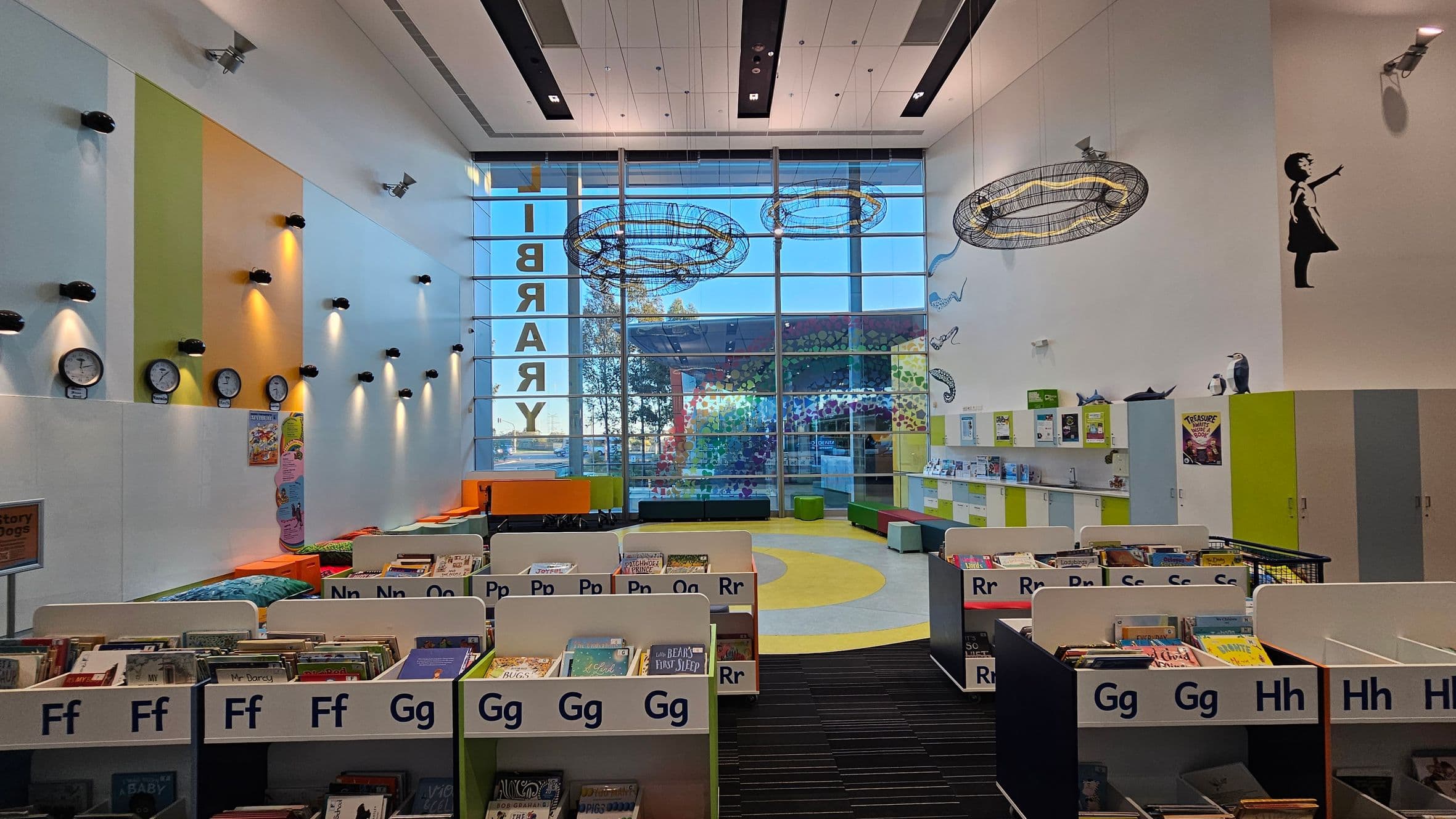 A modern library interior with a high ceiling and large glass windows at the back. The left wall features colourful panels with multiple clocks and spotlights. In the centre, there are bookshelves labelled with letters, filled with books. The right wall has a silhouette of a girl reaching out, and shelves with books and decorations. The floor is a mix of carpet and circular patterns, and there are colourful seating areas near the windows.