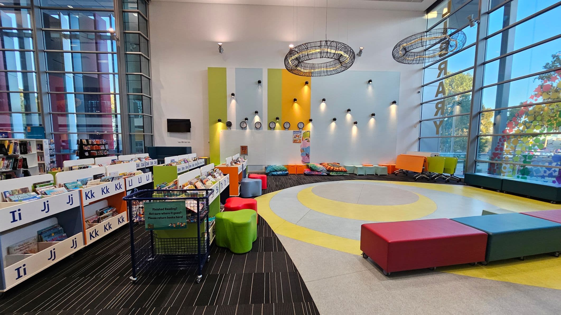 A spacious library area with large windows on the left and right, allowing natural light to flood in. On the left, there are shelves filled with books, organised alphabetically. In the centre, colourful seating options, including red, green, and blue stools, are arranged around a circular carpet with yellow and grey patterns. The back wall features clocks and spotlights, with additional seating and tables in orange and green. The floor is a mix of striped carpet and smooth tiles.