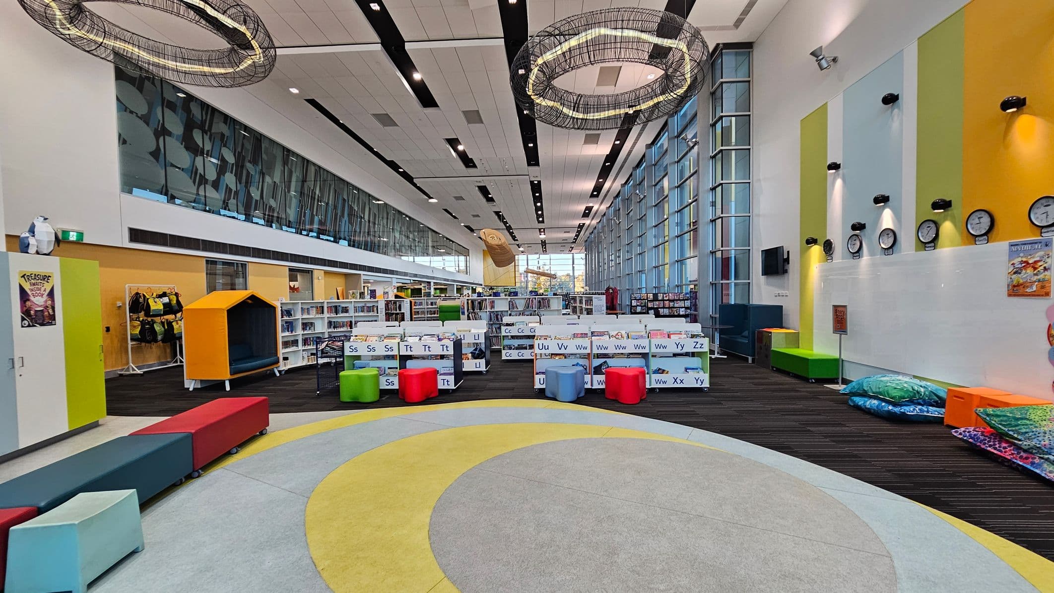 A spacious library interior with a high ceiling featuring circular light fixtures. The floor has a circular pattern in grey and yellow. On the left, there are colourful benches and a small house-shaped reading nook. In the centre, low bookshelves are organised alphabetically with colourful stools nearby. The right wall has vertical stripes in yellow and blue, with several clocks and a row of cushions on the floor. Large windows line the right side, allowing natural light to fill the space.