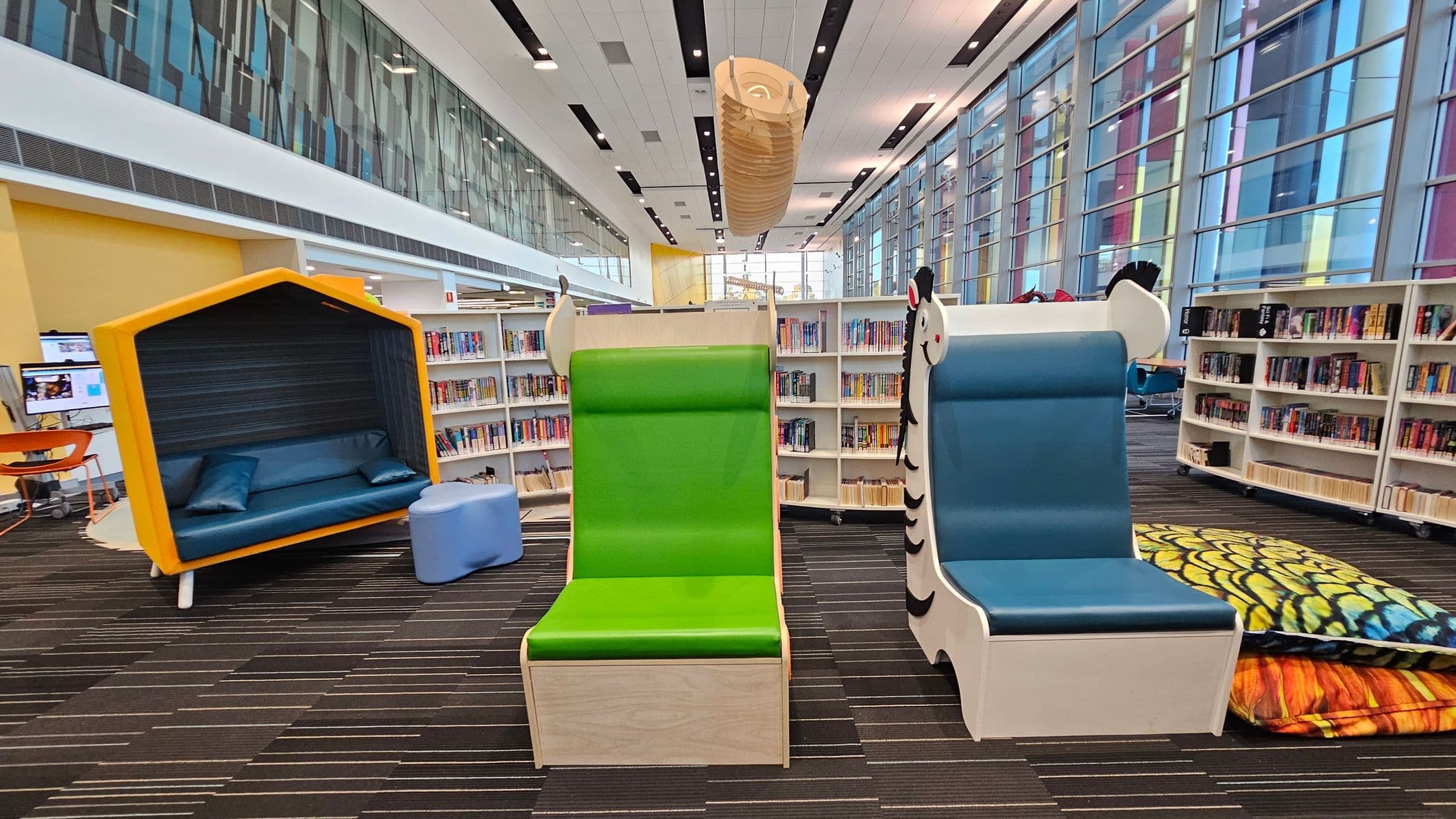 A modern library interior with colourful furniture. On the left, there's a yellow, house-shaped reading nook with a blue cushioned seat and pillows. In the centre, a green chair with a wooden base is next to a blue chair with a zebra design. On the right, large, patterned cushions are stacked. The background features bookshelves filled with books and a wall of large windows with vertical coloured panels. The ceiling is white with black accents and a large, cylindrical light fixture.