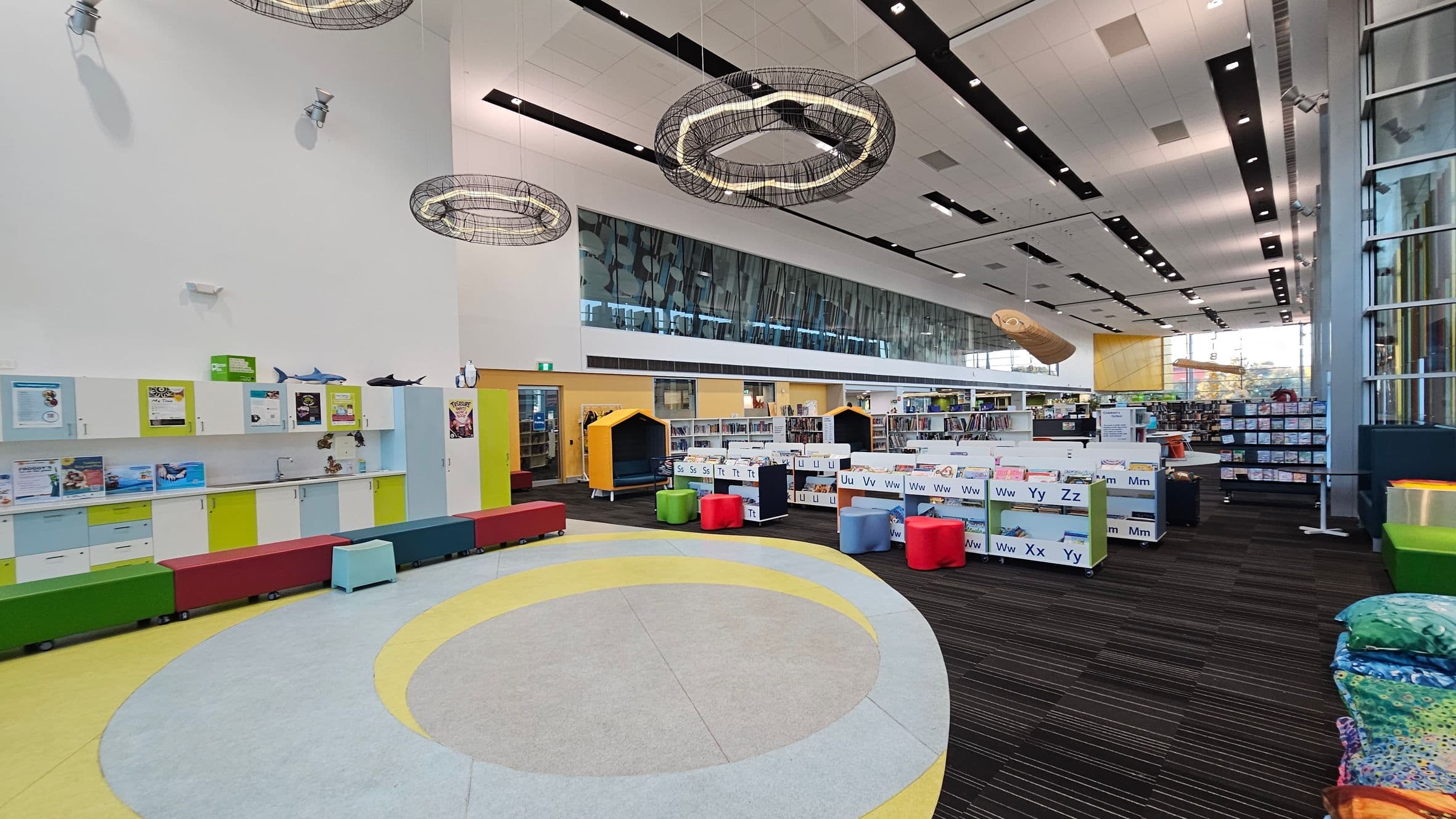 A spacious library interior with a high ceiling featuring modern light fixtures. On the left, there are colourful cabinets and a long bench with various hues. In the centre, circular patterns decorate the floor, surrounded by small, colourful stools and alphabet-labelled book bins. The right side has large windows and more bookshelves. The floor transitions from a light circular pattern to dark striped carpeting.