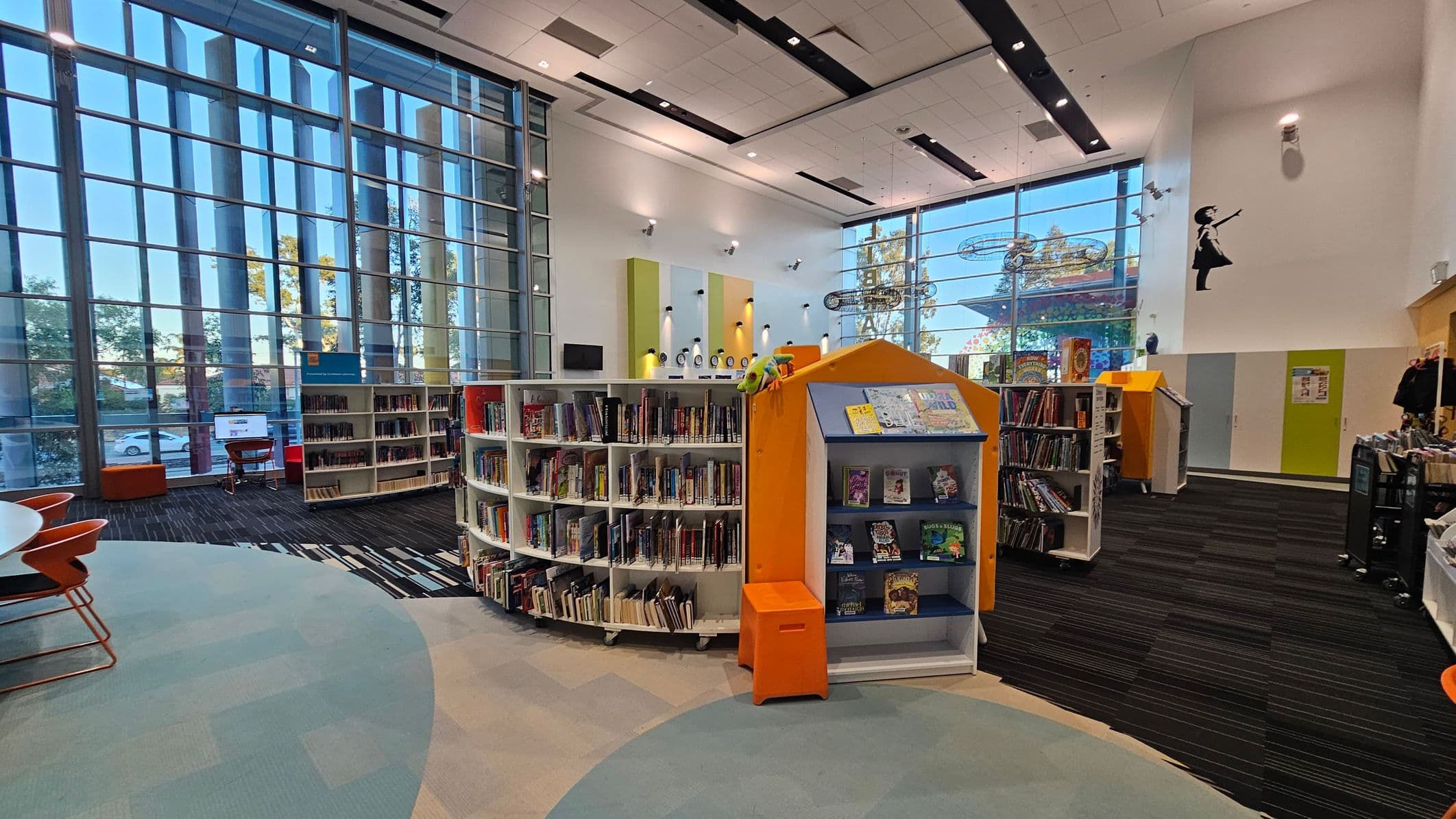 A modern library interior with large floor-to-ceiling windows on the left, allowing natural light to illuminate the space. In the centre, there are curved bookshelves filled with books, and a small orange stool in front of a display shelf. The floor has a mix of light blue and grey carpeting. On the right, more bookshelves and a wall with colourful panels. The ceiling is high with recessed lighting, and a silhouette of a girl with an umbrella is painted on the wall.