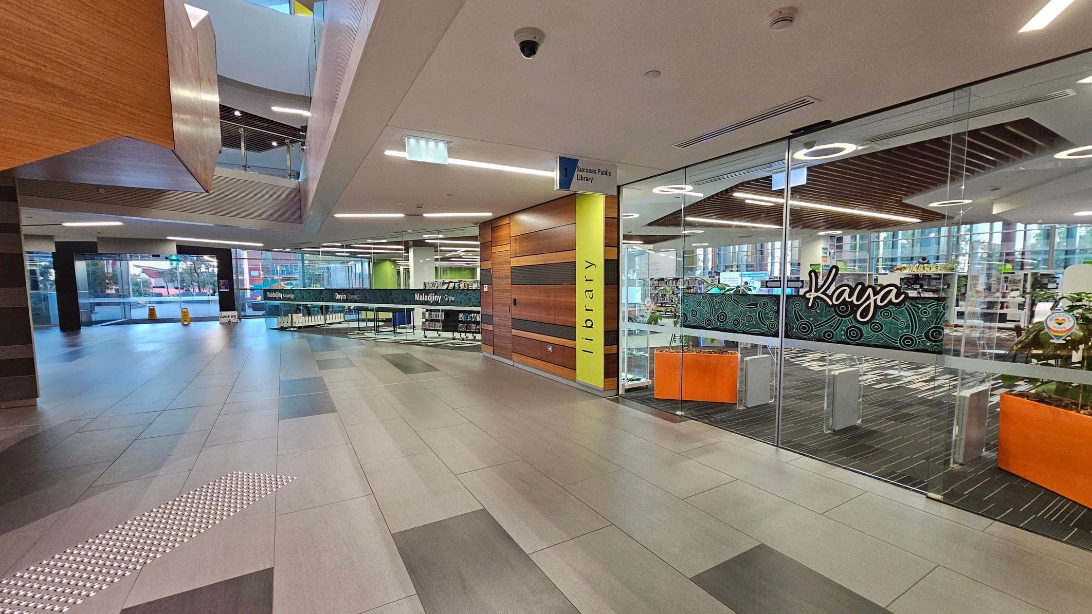 Modern interior with a spacious corridor. On the right, a glass-walled library entrance with "Kaya" written on it, featuring wooden panelling and a yellow column marked "library". Inside, bookshelves and a patterned carpet are visible. The left side has large windows and a tiled floor with a tactile strip. The ceiling has recessed lighting and a security camera. In the background, a sign for "Success Public Library" and a view of the outside through the windows.
