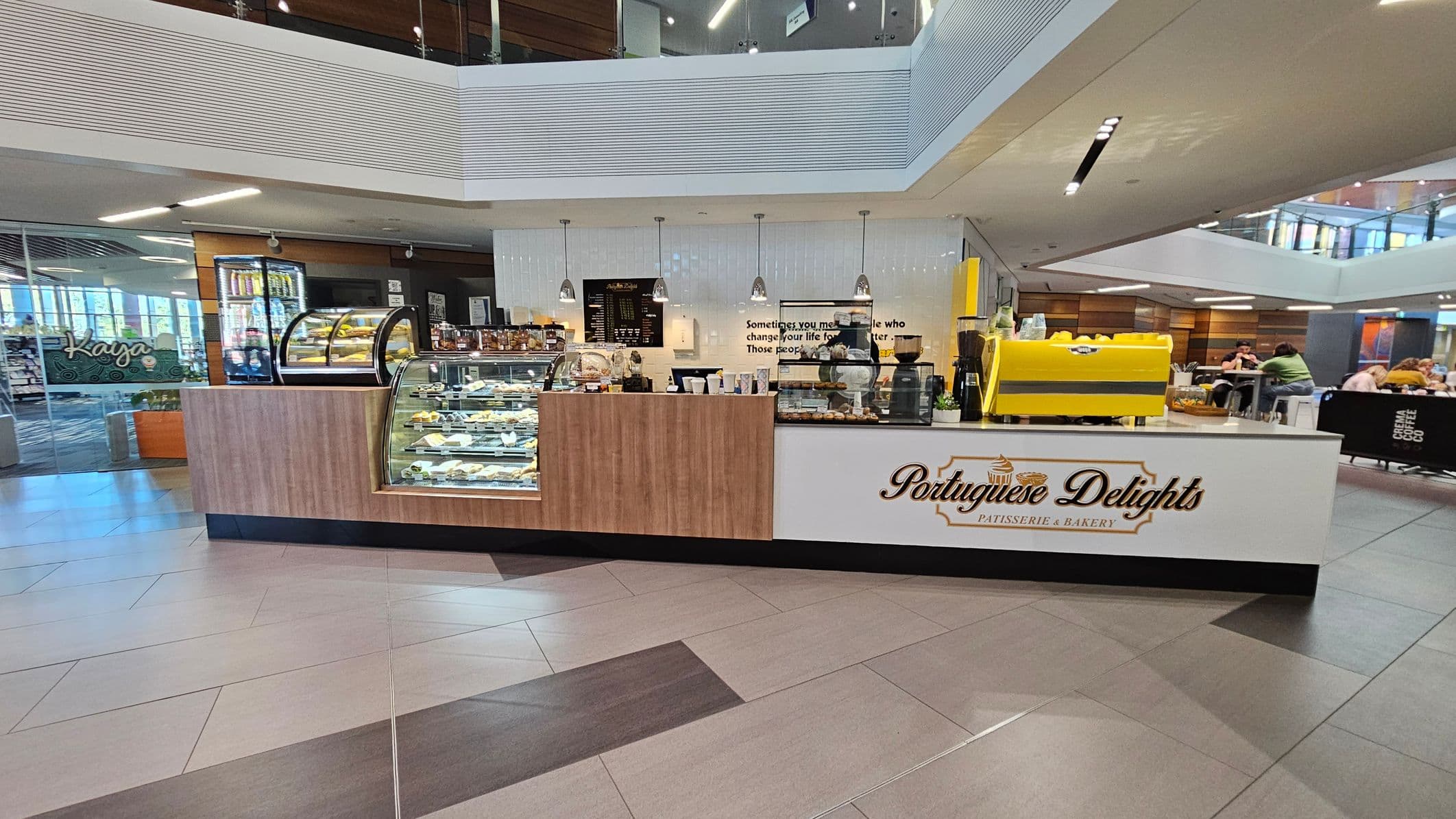 A bakery counter with a sign reading "Portuguese Delights Patisserie & Bakery" is in the centre. The counter displays various pastries in glass cases. On the right, a bright yellow coffee machine is visible. The background features a modern interior with tables and people seated. The ceiling has recessed lighting, and the floor is tiled in a mix of light and dark shades. On the left, there's a glass door with the word "Kaya" visible.