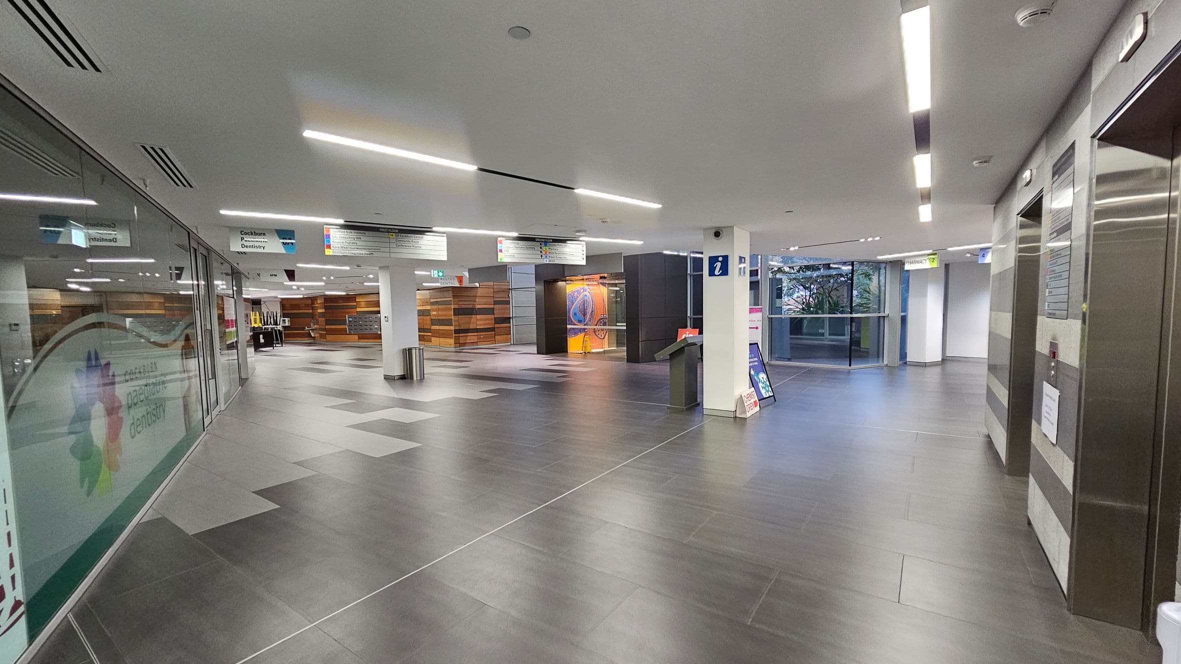A modern indoor space with grey tiled flooring and a white ceiling featuring recessed lighting. On the left, there is a glass wall with signage for a paediatric dentistry. In the centre, a pillar with an information sign and a digital kiosk is visible. The background shows a wooden panelled wall and a seating area. On the right, there are elevators with metallic doors. A window at the far end provides natural light, and there are various directional signs hanging from the ceiling.