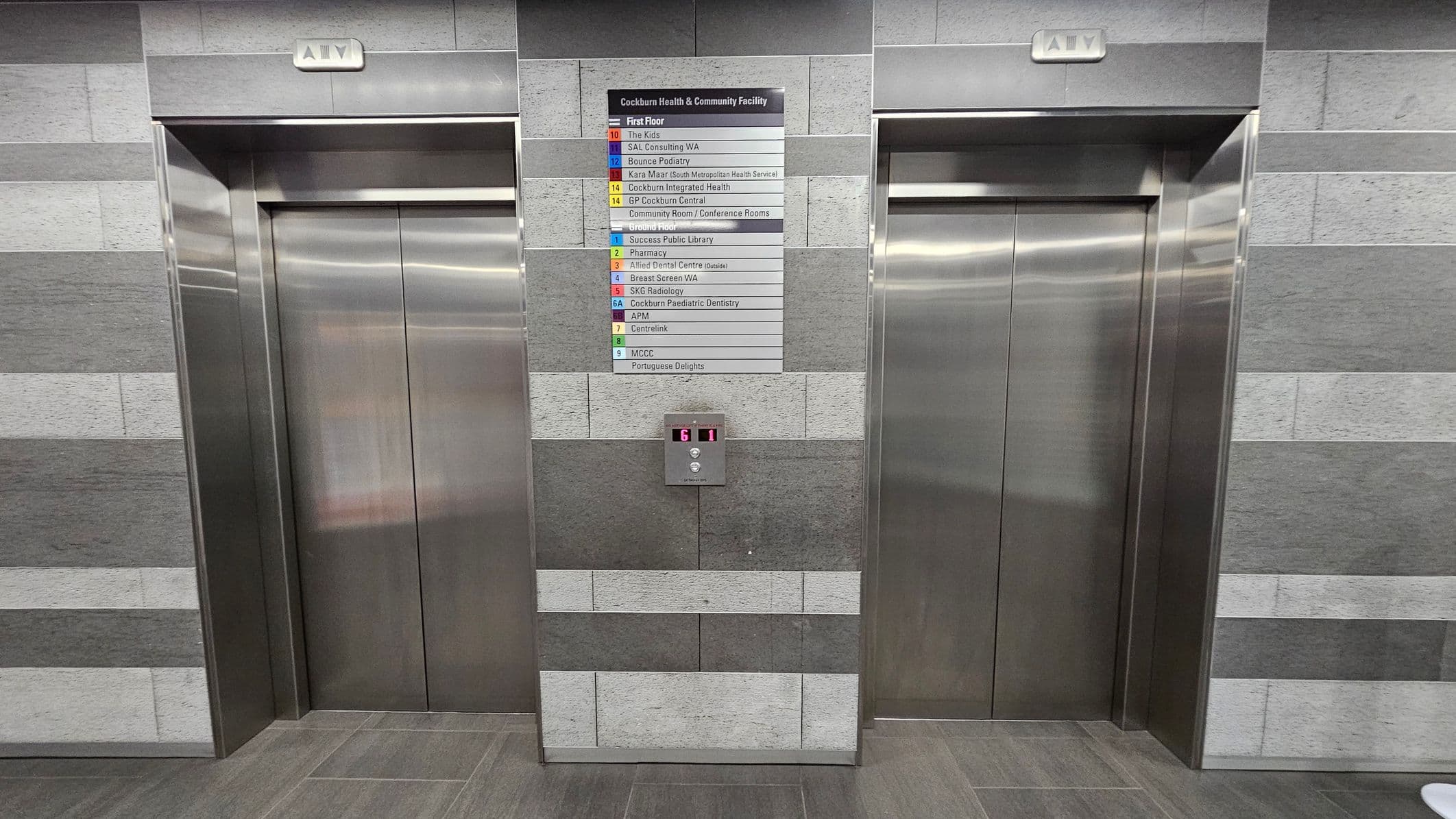 Two stainless steel lift doors are set into a wall with a grey, horizontally striped tile pattern. Above each lift is a sign with the word "LIFT". Between the lifts, a directory lists various facilities and their corresponding floor numbers. Below the directory, a digital display shows the number "G" and an arrow pointing up. The floor is tiled in a matching grey tone.
