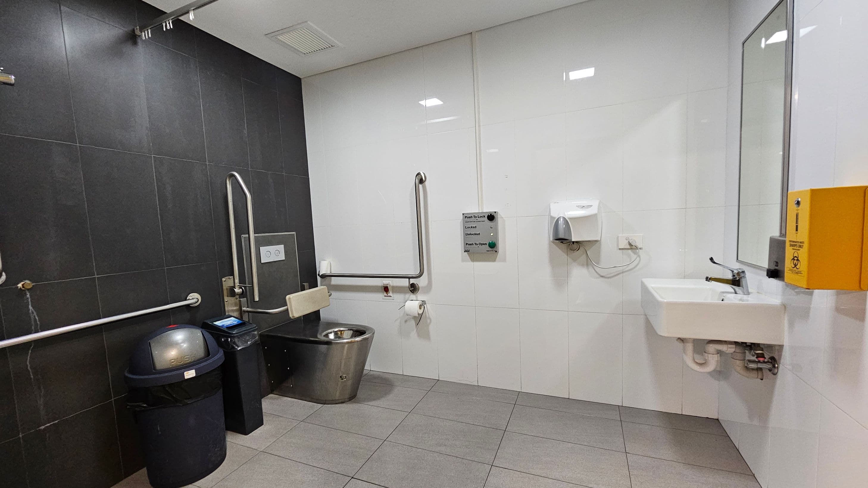 A public restroom with accessibility features. On the left, there is a black tiled wall with a stainless steel toilet equipped with support rails. A black waste bin and a small black box are nearby. The right side has white tiles, a white sink with a mirror above, and a yellow box mounted on the wall. A soap dispenser is also present. The floor is grey tiled, and the ceiling is white with recessed lighting.