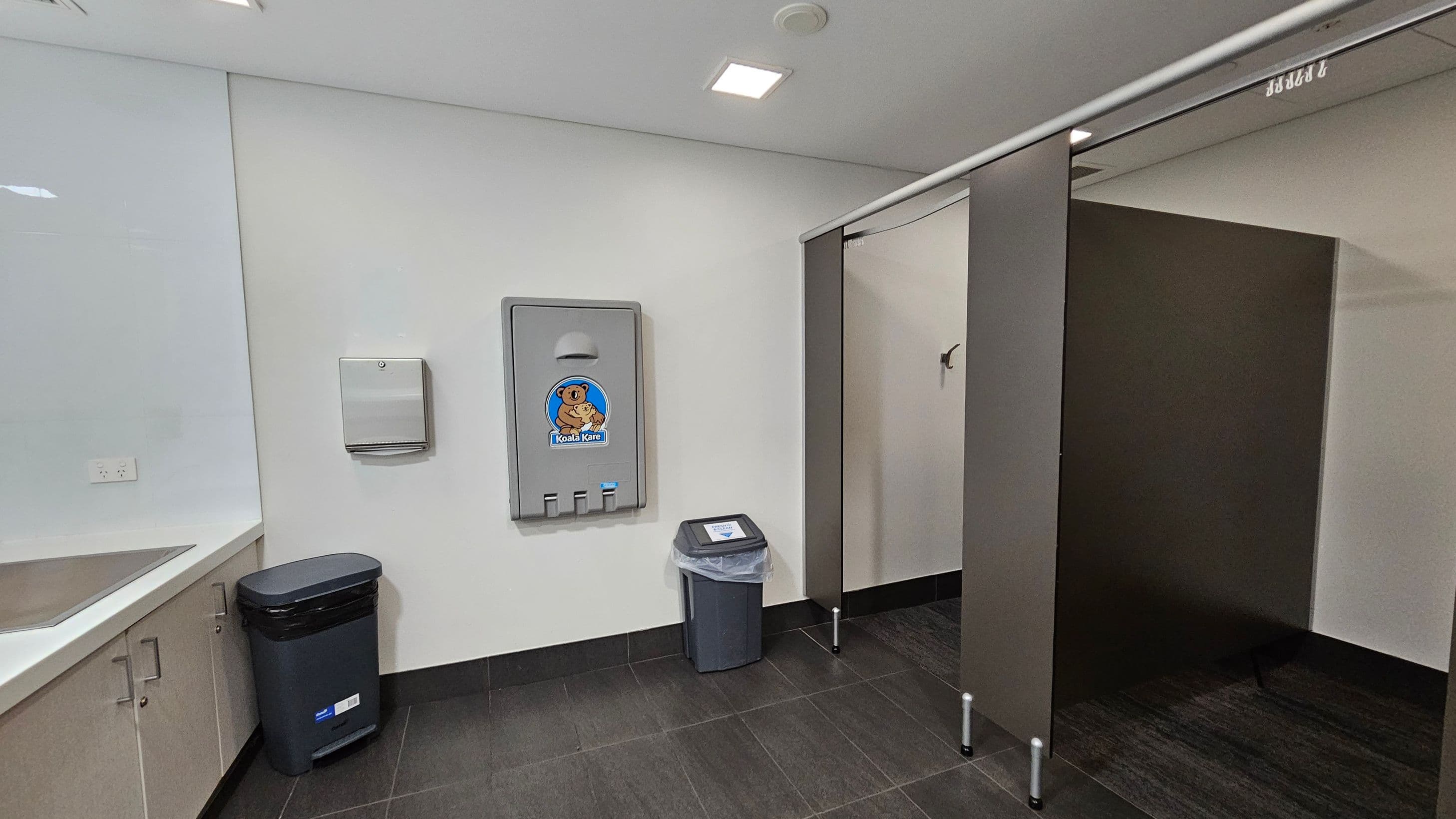 A public restroom with a baby changing station on the left wall, featuring a cartoon koala logo. Below it is a small bin with a liner. To the left, a countertop with a sink and wooden cabinets underneath. A paper towel dispenser is mounted on the wall above a larger bin. On the right, there are dark grey stall dividers with a partially open door. The floor is covered with dark tiles, and the ceiling is white with recessed lighting.