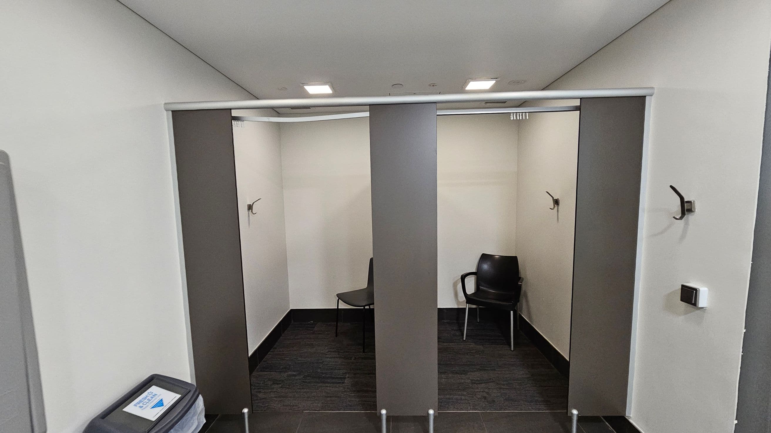 Two changing cubicles with grey curtains partially open. Each cubicle has a chair and a wall hook. The floor is dark grey, and the walls are light-coloured. A waste bin with a sign is on the left. The ceiling is white with recessed lighting.