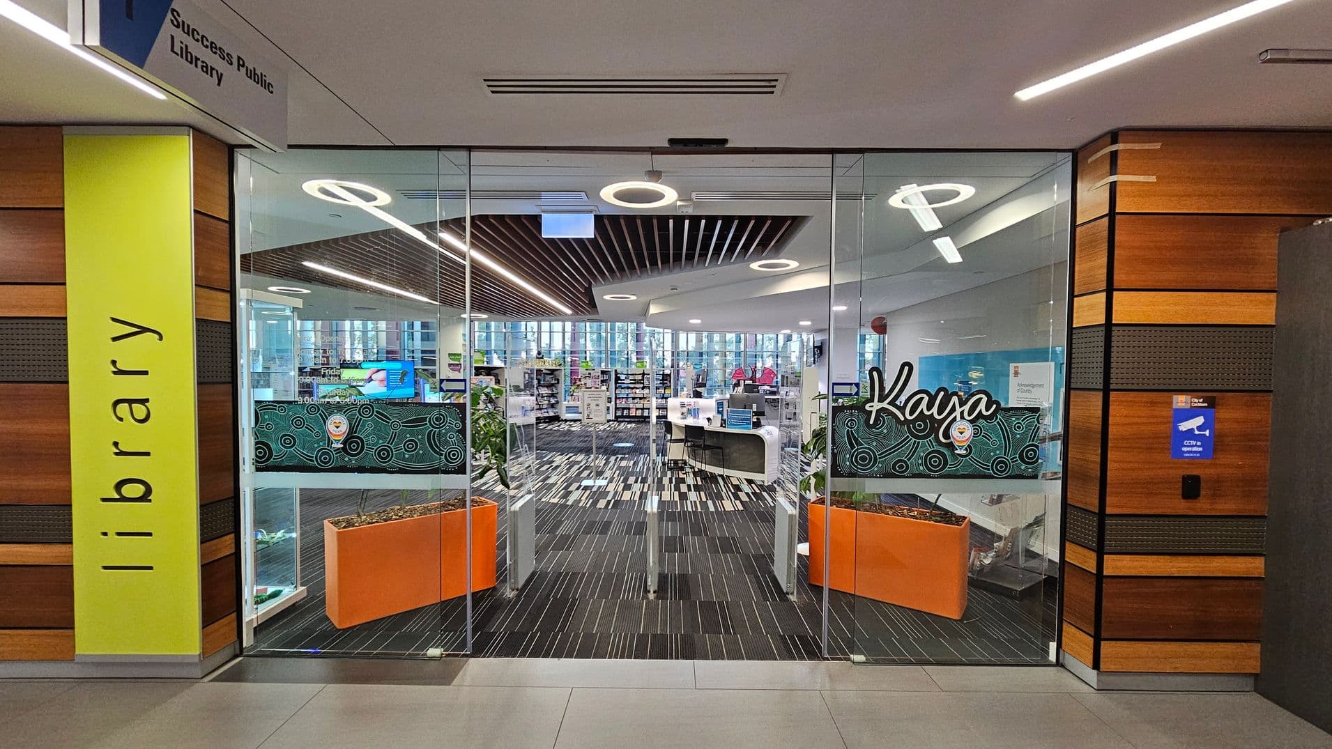 Glass entrance to a library with a bright yellow panel on the left displaying the word "library" vertically. Above, a sign reads "Success Public Library." The interior features a modern design with striped flooring and circular ceiling lights. Two orange planters are visible near the entrance. Bookshelves and a reception desk are in the background. The word "Kaya" is displayed on the glass doors with a decorative pattern. Walls are lined with wood panelling.