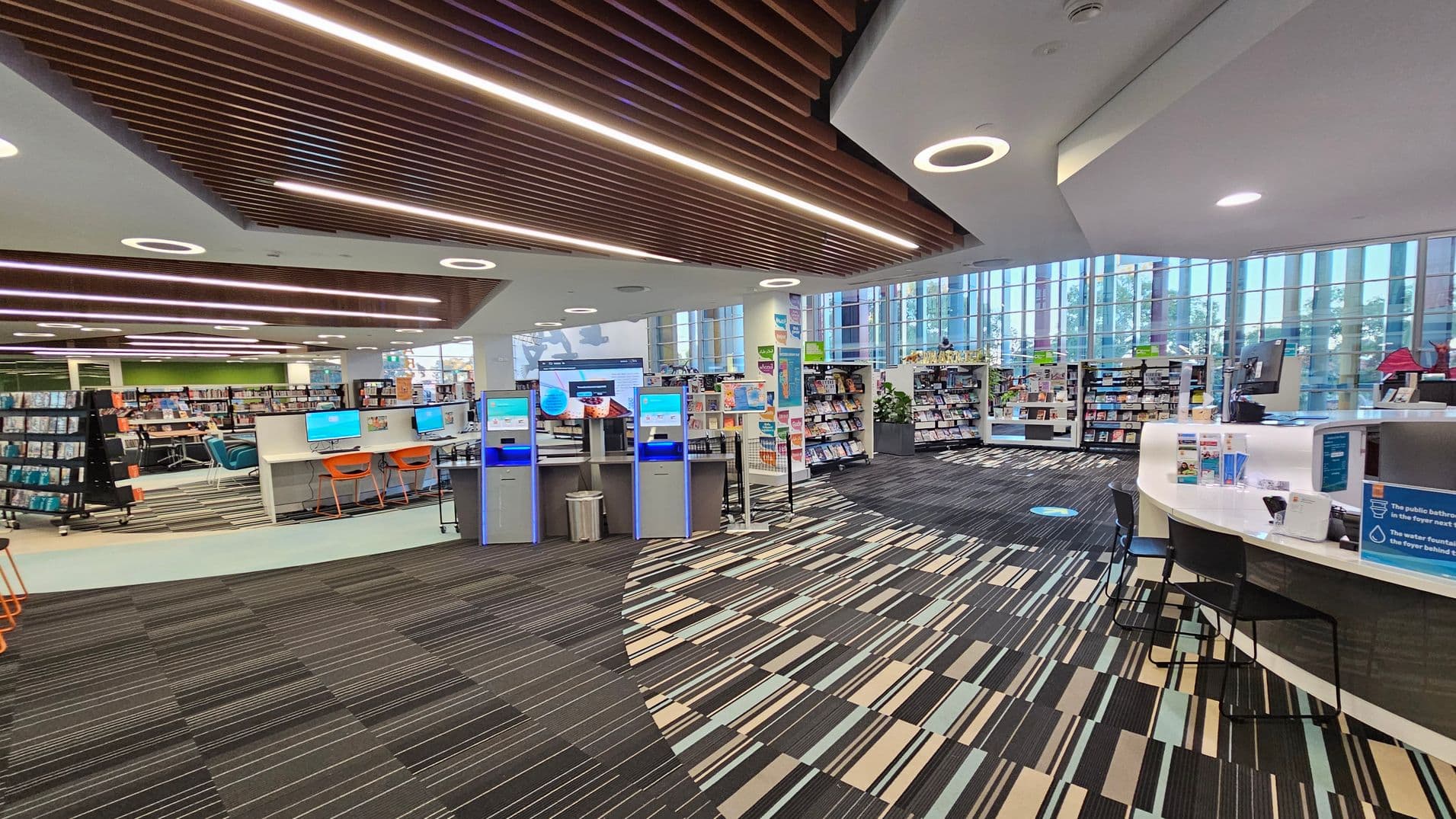 A modern library interior with a striped carpeted floor. On the left, there are bookshelves and a row of computers with orange chairs. In the centre, several self-service kiosks are visible. On the right, a curved desk with informational brochures and a computer. The background features large windows allowing natural light, and more bookshelves. The ceiling has wooden slats and circular lights.