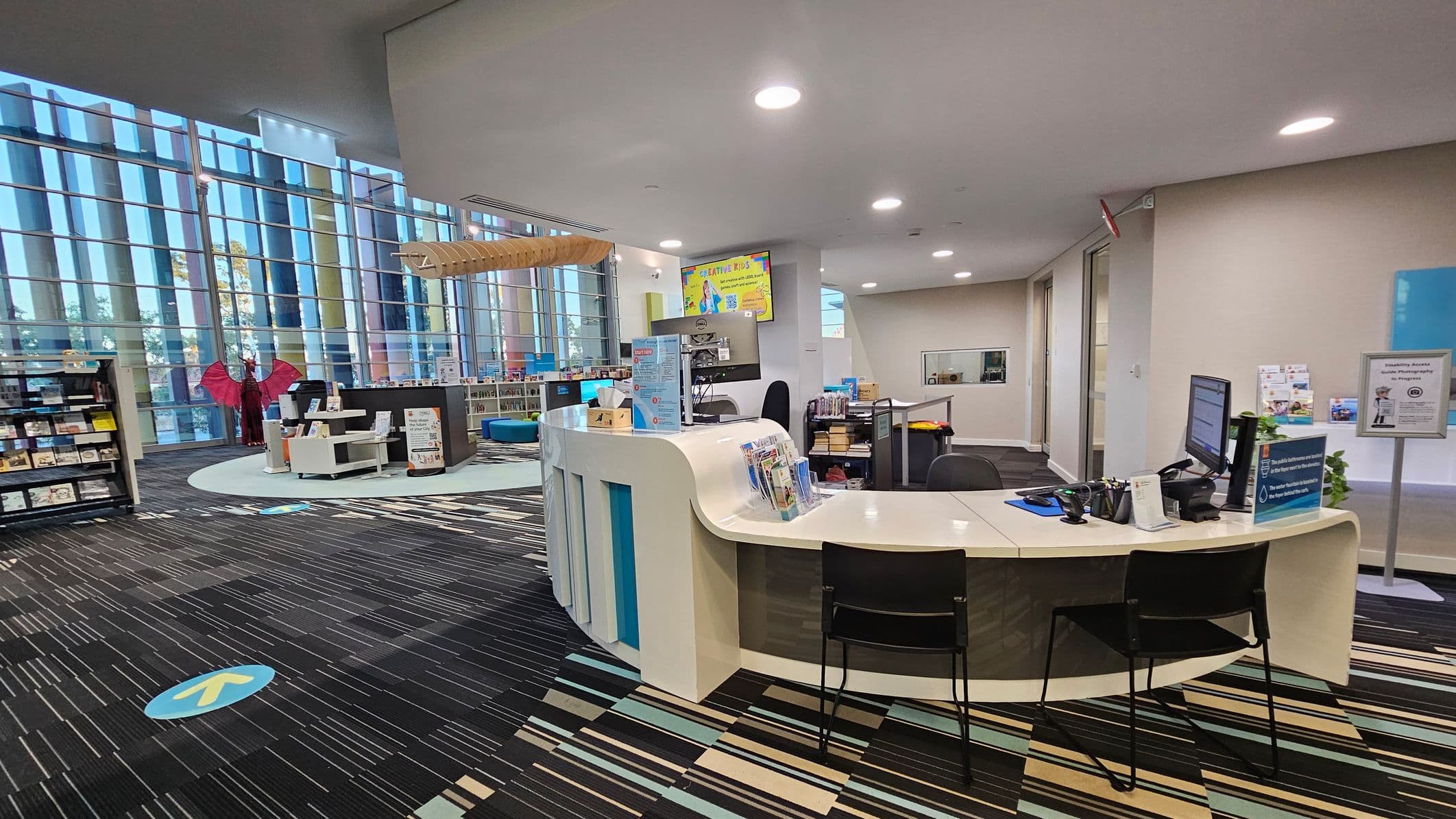A modern library interior with a curved reception desk in the foreground, equipped with a computer and informational brochures. To the left, there are bookshelves and a mannequin with red wings. The background features large glass windows allowing natural light, and a colourful carpet with a striped pattern covers the floor. A digital screen displays a sign above the desk area. The ceiling is white with recessed lighting.