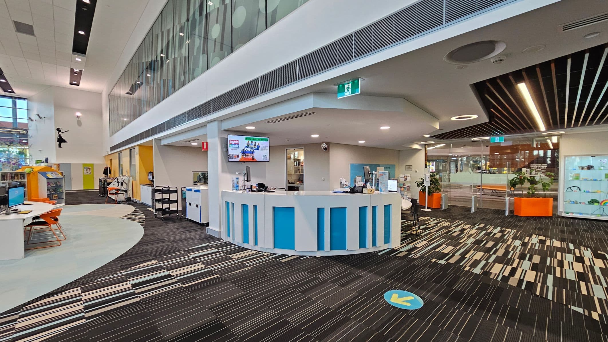 A modern library interior with a curved reception desk in the centre, featuring blue panels. To the left, there are computer stations with orange chairs and a colourful carpet. The right side has a glass wall with orange planters and display shelves. The ceiling has recessed lighting and a mix of white and wooden slats. The floor is patterned with stripes in various colours. An exit sign is visible above the reception area.