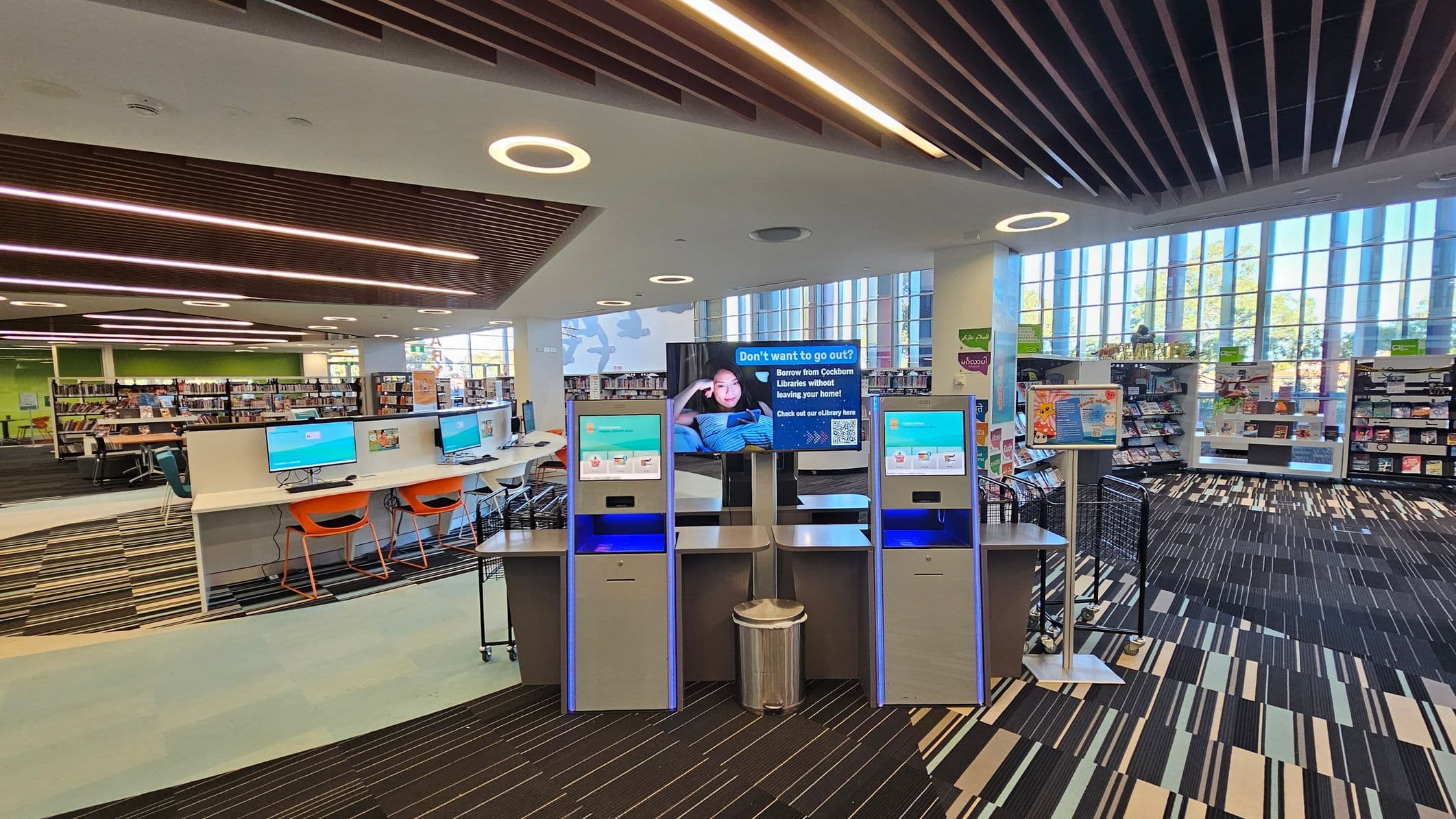 A modern library interior with a striped carpeted floor and a ceiling featuring circular lights and wooden slats. In the centre, there are self-service kiosks with blue screens and a digital display promoting library services. To the left, a row of computers with orange chairs is visible. The background shows bookshelves filled with books and large windows allowing natural light to enter.