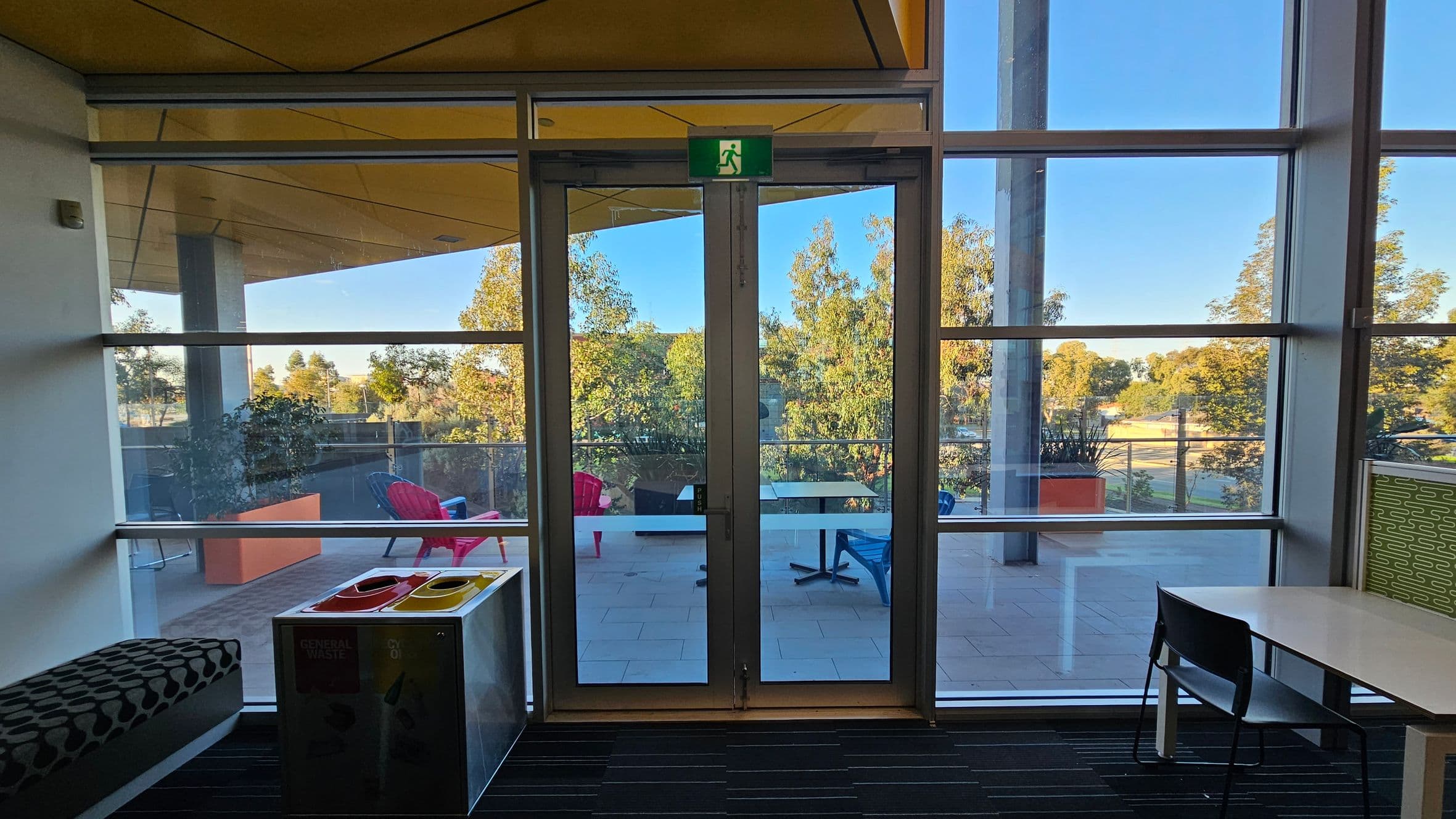 Glass doors with an emergency exit sign lead to an outdoor area. Outside, there are red and blue chairs and tables on a tiled floor, with trees visible in the background. Inside, on the left, a patterned bench and a waste bin with recycling compartments are present. On the right, a white table with a black chair is positioned near the window. The ceiling is yellow, and the floor has a striped pattern.