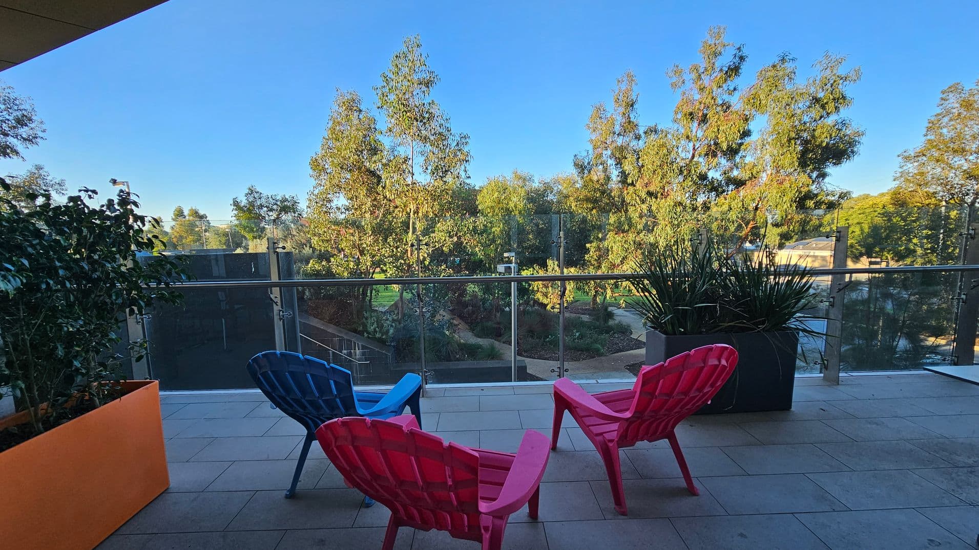 A balcony with two colourful chairs, one pink and one blue, facing a glass railing. On the left, there's a large orange planter with green foliage. On the right, a dark planter holds tall, spiky plants. Beyond the railing, a garden with trees and pathways is visible. The floor is tiled, and the ceiling is not visible.