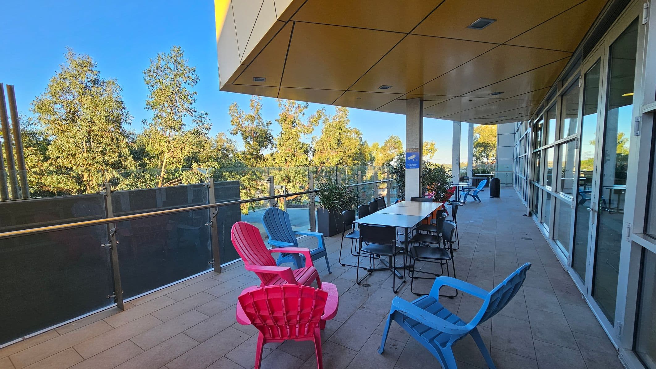 A spacious outdoor terrace with a tiled floor. On the left, there are colourful chairs in pink and blue, and a glass railing with trees visible beyond it. In the middle, a long table with several chairs is set up, accompanied by potted plants. On the right, large glass windows reflect the scene. The ceiling is covered with yellow panels, and there are columns supporting the structure.