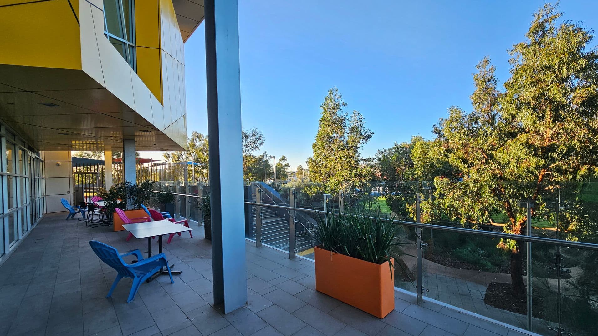 A terrace with a tiled floor and a glass railing on the right, overlooking a garden with trees. On the left, a building with yellow and white panels extends overhead. Several colourful chairs and tables are arranged along the terrace, with a few potted plants nearby. Stairs descend from the terrace to the garden area below.