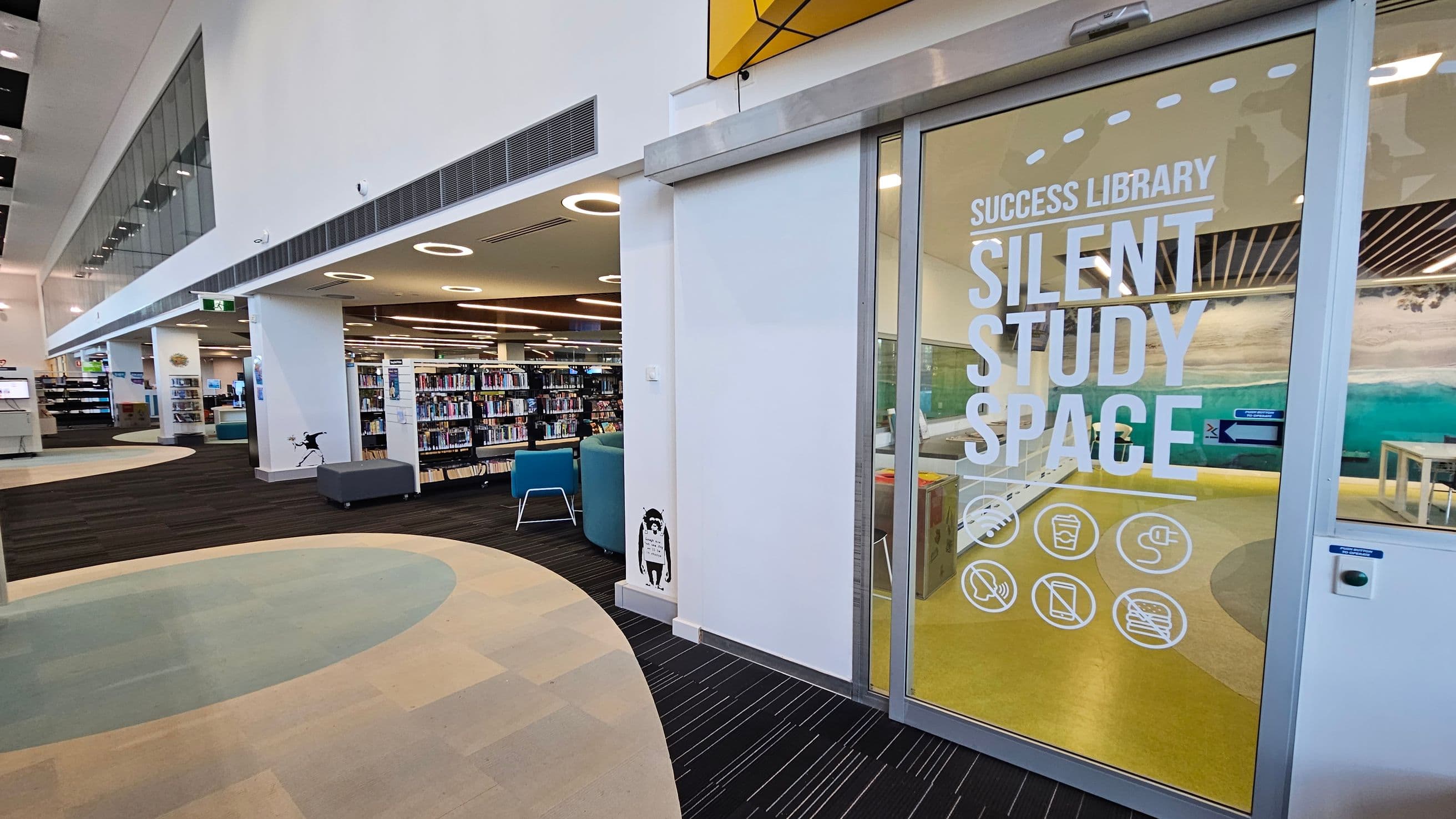 A modern library interior with a focus on a glass door on the right, labelled "Success Library Silent Study Space" with icons below. Inside, there are tables and chairs on a yellow floor. To the left, the library features bookshelves, blue seating, and a patterned carpet with circular designs. The ceiling is high with circular lights and air vents. A small black-and-white figure is painted on the wall near the door.
