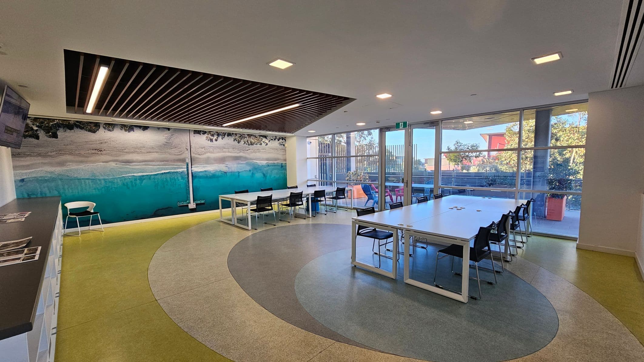 A spacious room with a large mural of a beach scene on the left wall. The floor has a circular pattern in shades of green and grey. In the centre, there are white tables with black chairs arranged around them. The right side features large windows with a view of trees and a red building outside. The ceiling has recessed lighting and a section with wooden slats. A white chair and a counter with shelves are on the left.