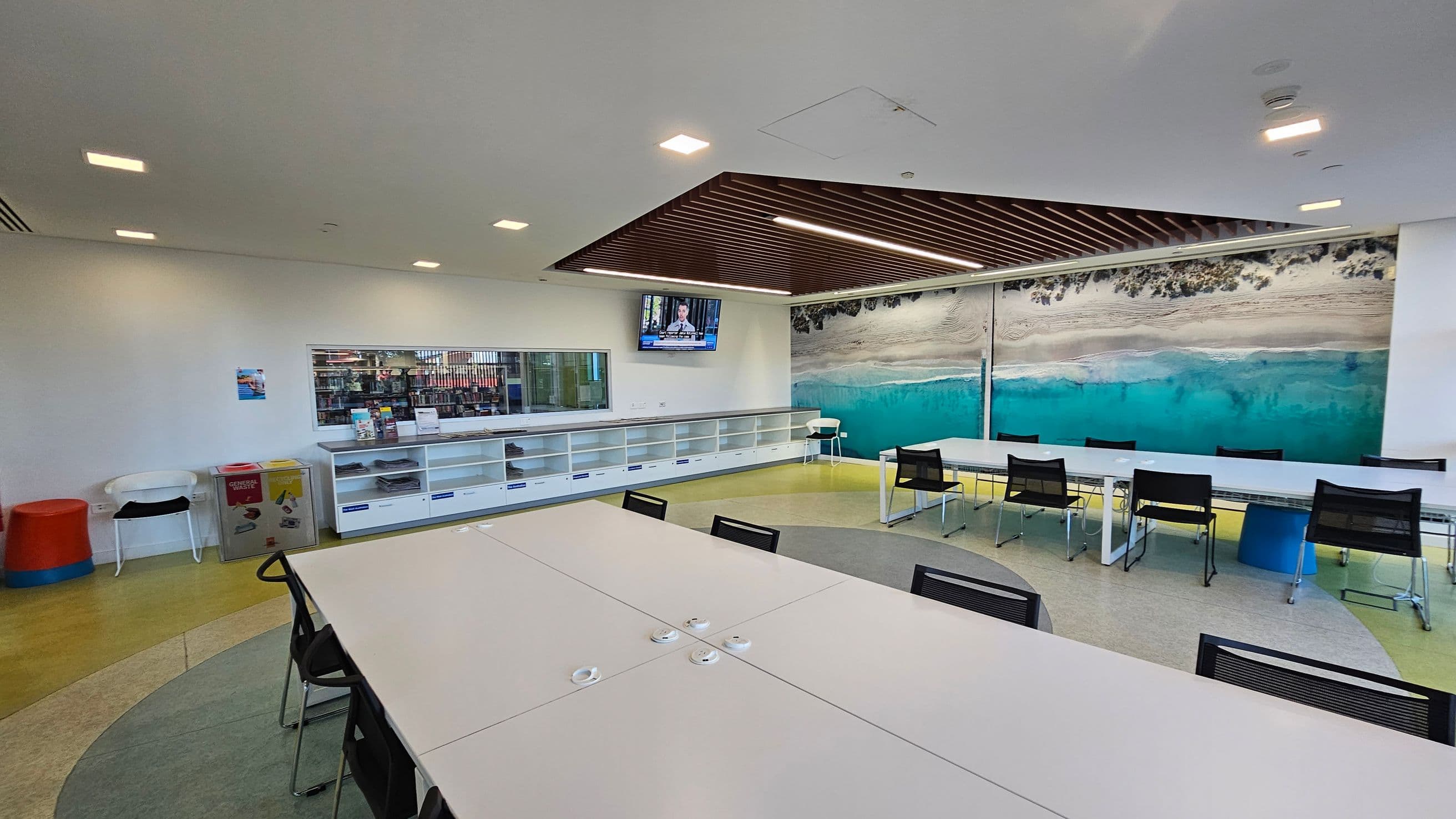 A modern room with a large table and black chairs in the foreground. The floor has a circular pattern with shades of grey and yellow. On the left, there are colourful bins and a white chair. The back wall features a large mural of a beach scene with turquoise water. Above, a television is mounted on the wall, and a wooden slatted ceiling section adds texture. Shelving units line the back wall, and a window reveals a glimpse of a library area.