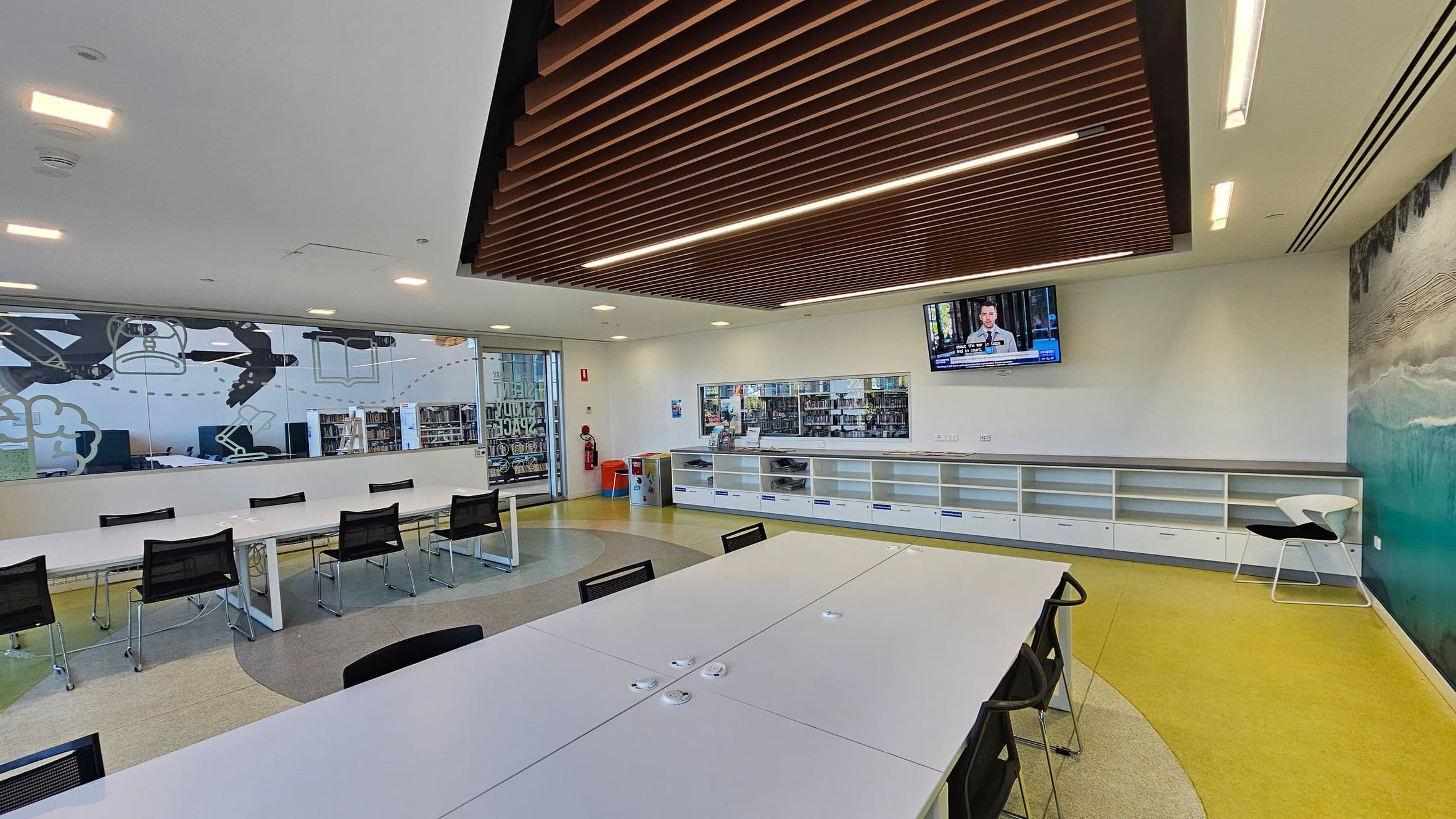 A modern room with a long white table and black chairs in the centre. The left wall features large windows with abstract designs and a view of bookshelves. The right wall has a mural with green and blue tones. A television is mounted on the wall above white cabinets. The ceiling has wooden slats, and the floor is a mix of yellow and grey. A fire extinguisher and a small bin are near the glass door on the left.