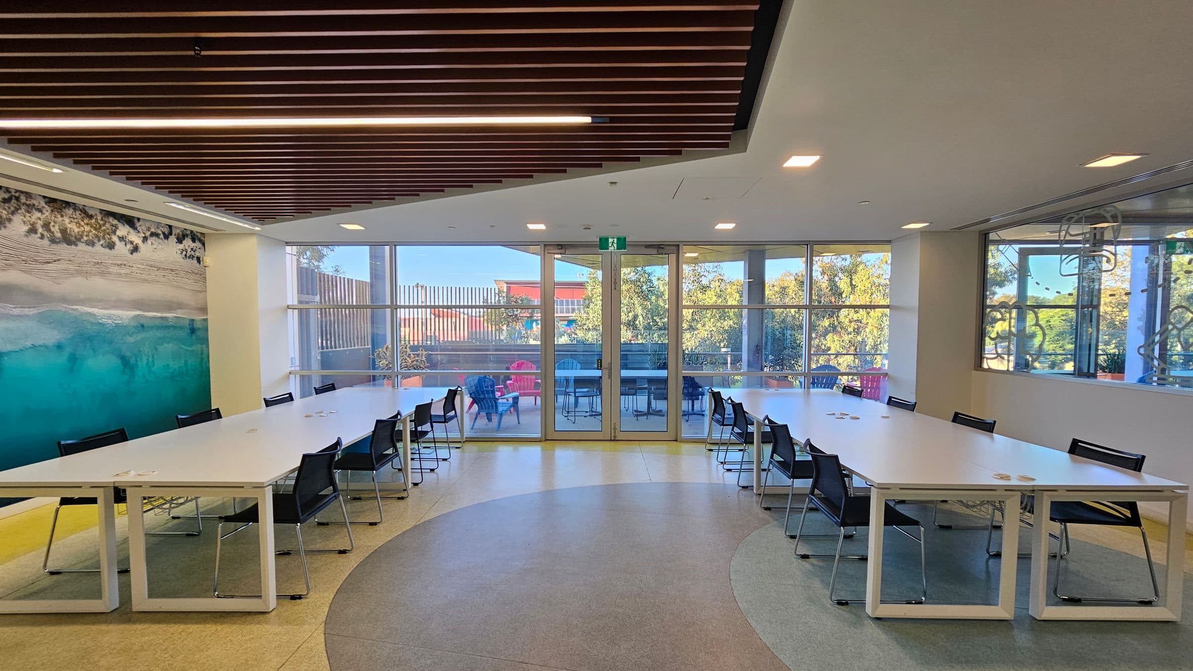 A modern meeting room with a large U-shaped white table surrounded by black chairs. On the left wall, there's a mural of a beach scene with sand and turquoise water. The back wall features large glass doors and windows, leading to an outdoor area with tables and chairs. The ceiling has wooden slats with integrated lighting. The floor is a mix of light and dark grey tiles, forming a circular pattern in the centre.