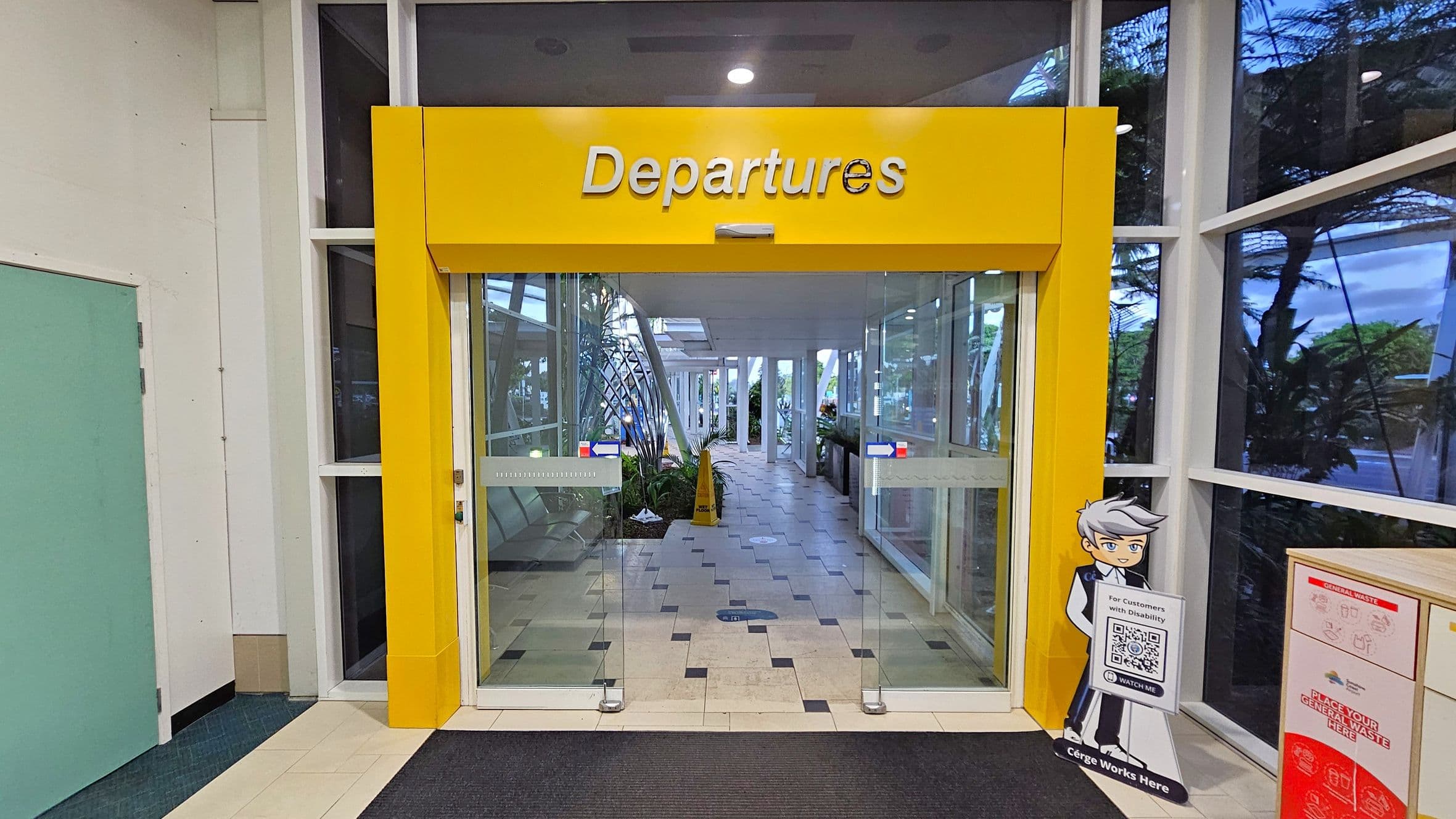 A yellow sign with "Departures" is above a set of glass sliding doors. To the right, there's a cartoon character cutout holding a sign with QR codes. The floor is tiled with a mix of light and dark squares, leading to a corridor with plants and large windows. On the left, there's a light blue door. The ceiling has recessed lighting.