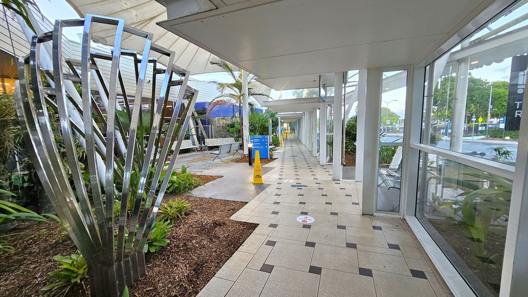 A covered walkway with a tiled floor featuring a geometric pattern. On the left, there's a metallic sculpture surrounded by plants and mulch. Further along, benches and a blue sign are visible. A yellow caution sign is placed on the floor. The right side has large glass windows, offering a view of trees and a street outside. The ceiling is flat and white, with a series of beams supporting the structure.