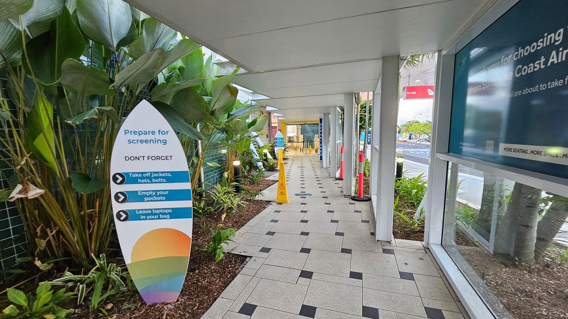 A covered walkway with a tiled floor leads to an entrance in the distance. On the left, there are large green plants and a sign reminding people to prepare for screening by removing jackets, hats, and belts, and emptying pockets. On the right, there is a glass wall with a blue sign. A yellow caution sign and red bollards are visible further along the path. The ceiling is white and flat.