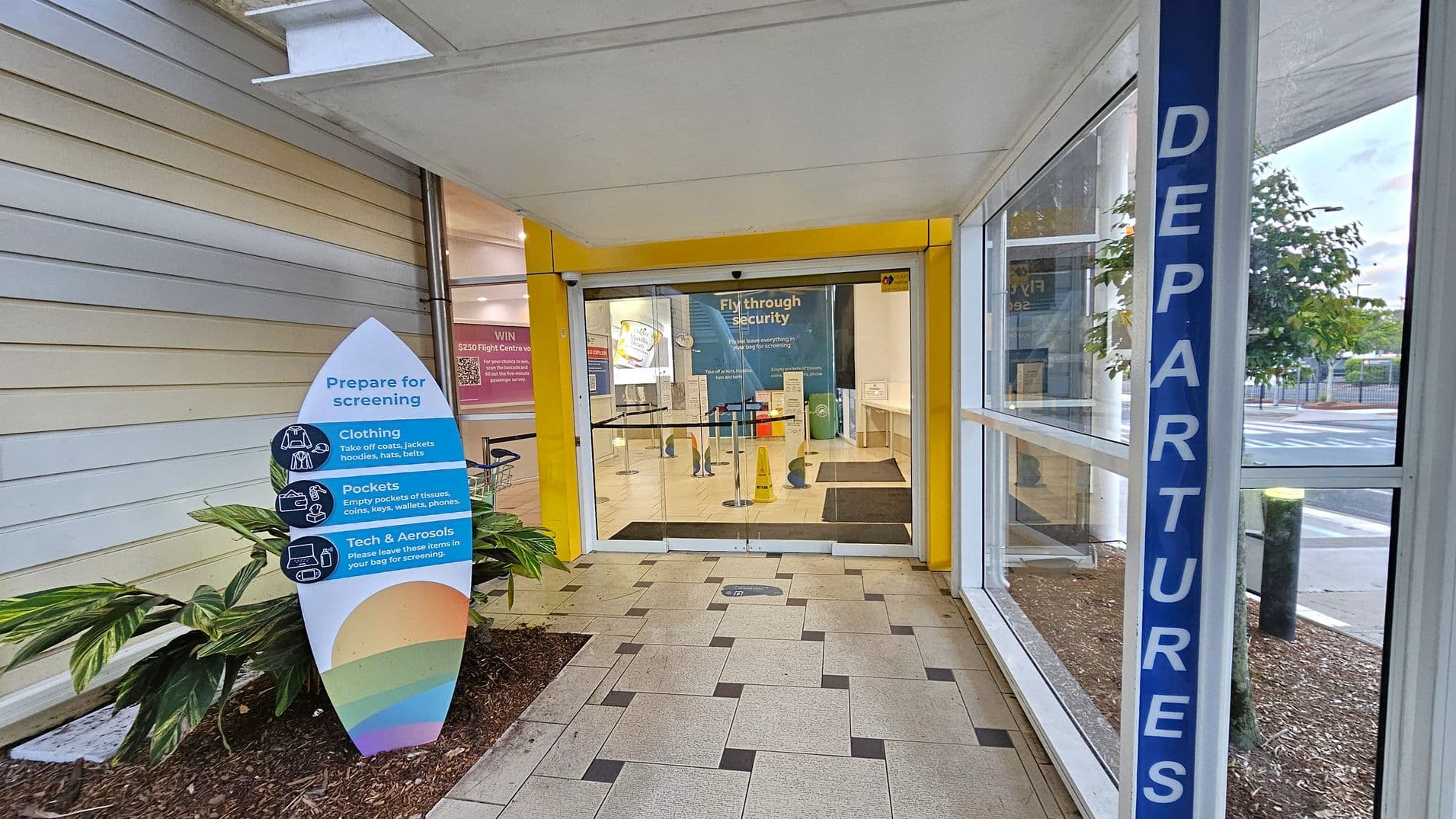 A corridor leading to a security screening area. On the left, there's a sign with instructions for preparing for screening, including clothing and pocket checks. Plants are visible at the base of the sign. The floor is tiled with a pattern of light and dark squares. On the right, a glass wall with a blue "DEPARTURES" sign. The entrance to the security area has yellow framing and a sign that reads "Fly through security." Outside, trees and a road are visible through the glass.
