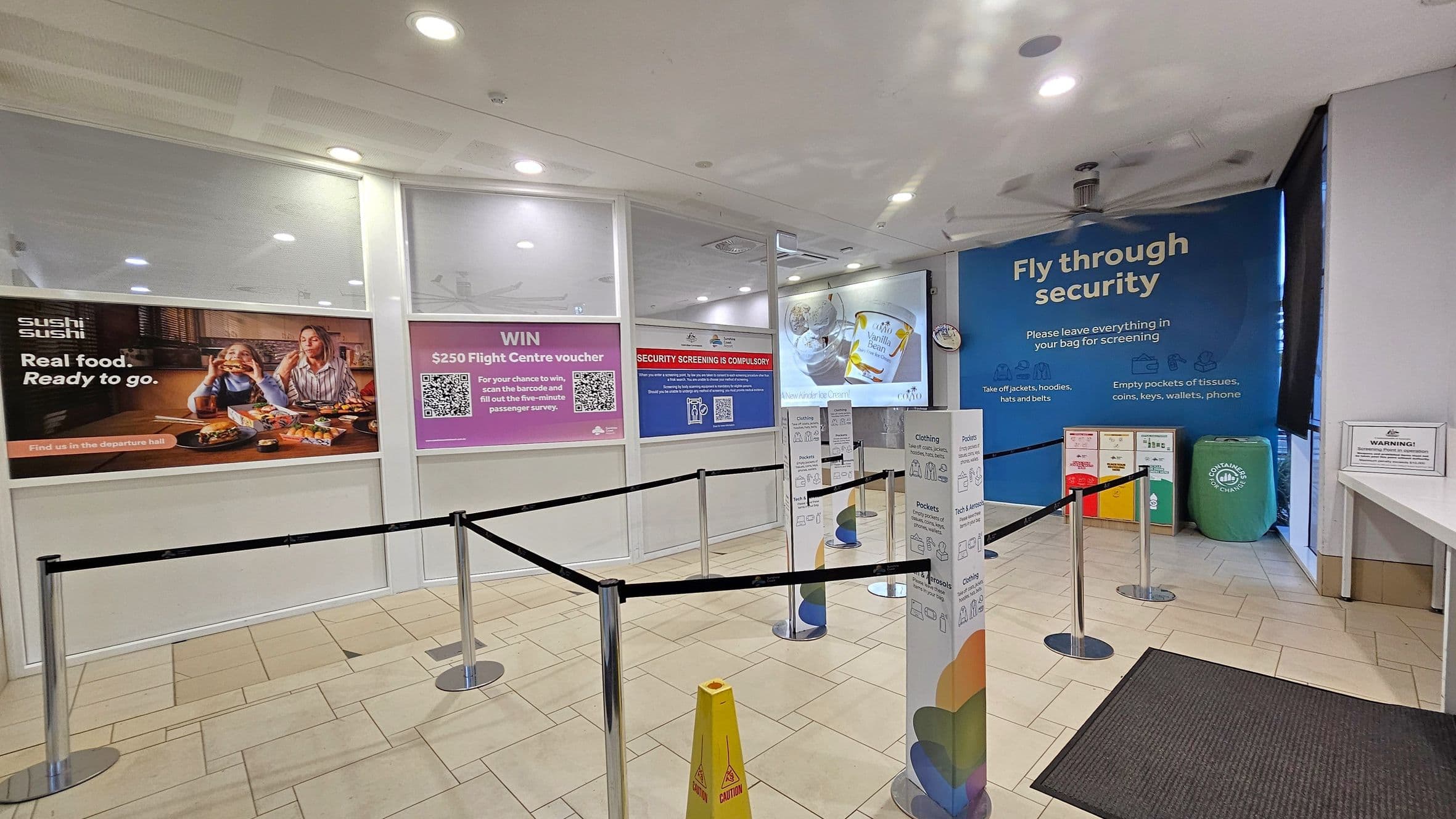 A security screening area with a tiled floor and a white ceiling with recessed lights. On the left, there are advertisements, including one for sushi and another offering a chance to win a voucher. In the centre, black retractable barriers guide the queue. On the right, a large blue sign instructs passengers to prepare for security checks. Recycling bins are visible in the background. A caution sign is on the floor near the entrance.
