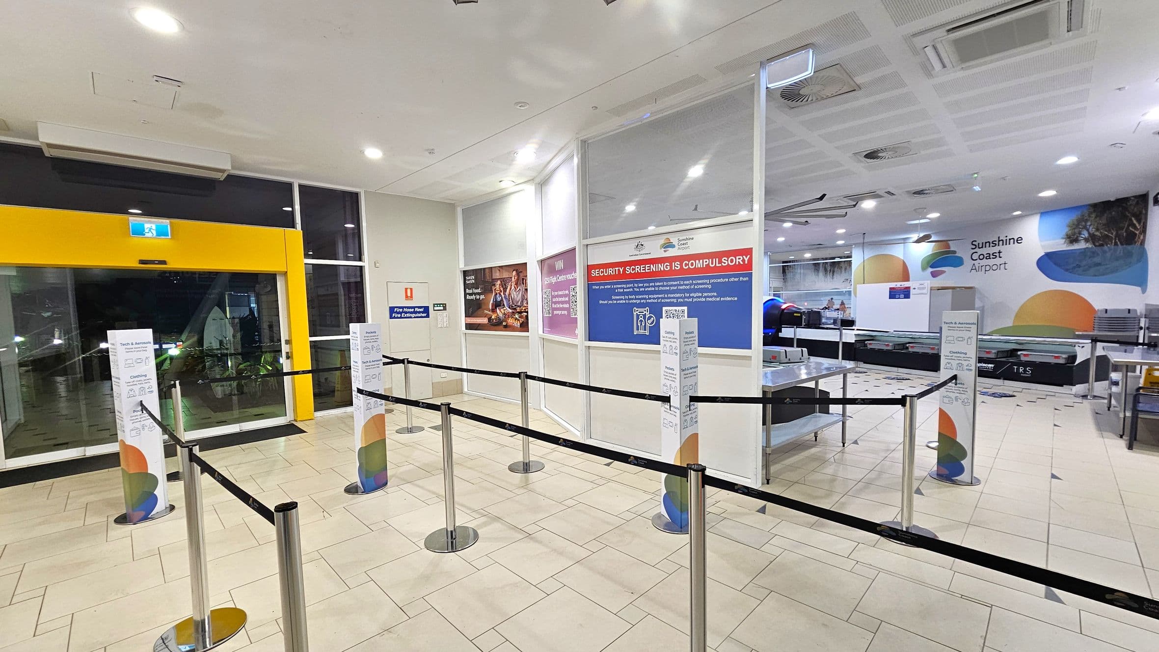 A spacious airport security area with a tiled floor. On the left, a yellow-framed glass exit door with an illuminated exit sign above. In the centre, retractable barriers guide passengers, with informational signs on pillars. On the right, a security screening area with a conveyor belt and a large wall sign reading "Sunshine Coast Airport." The ceiling has recessed lighting and air vents. Walls display various informational posters.