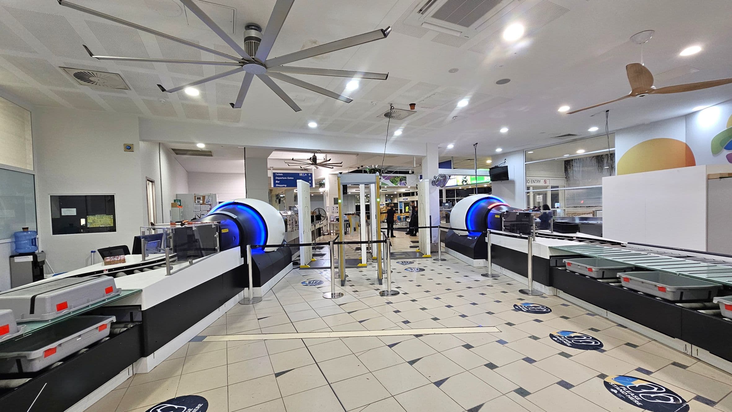Security checkpoint area with two large scanning machines on either side, each with a conveyor belt and grey trays. In the centre, there are metal detectors and a barrier with a sign above indicating directions. The floor is tiled with a pattern, and there are social distancing markers. The ceiling features large fans and recessed lighting. Walls have colourful abstract designs, and a water cooler is visible on the left.