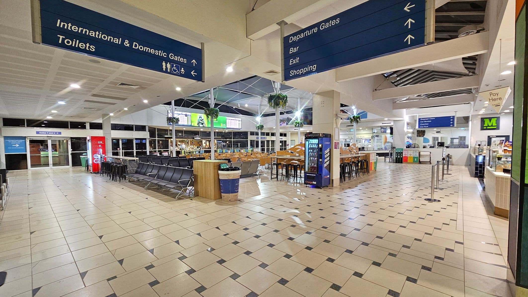 An airport terminal with a tiled floor and a high ceiling. On the left, there are signs for international and domestic gates and toilets, with a Coca-Cola vending machine nearby. In the centre, there are rows of black seating and a bar area with tables and chairs. On the right, a vending machine and a café counter with a "Pay Here" sign are visible. Overhead signs direct to departure gates, bar, exit, and shopping. Hanging plants are suspended from the ceiling.