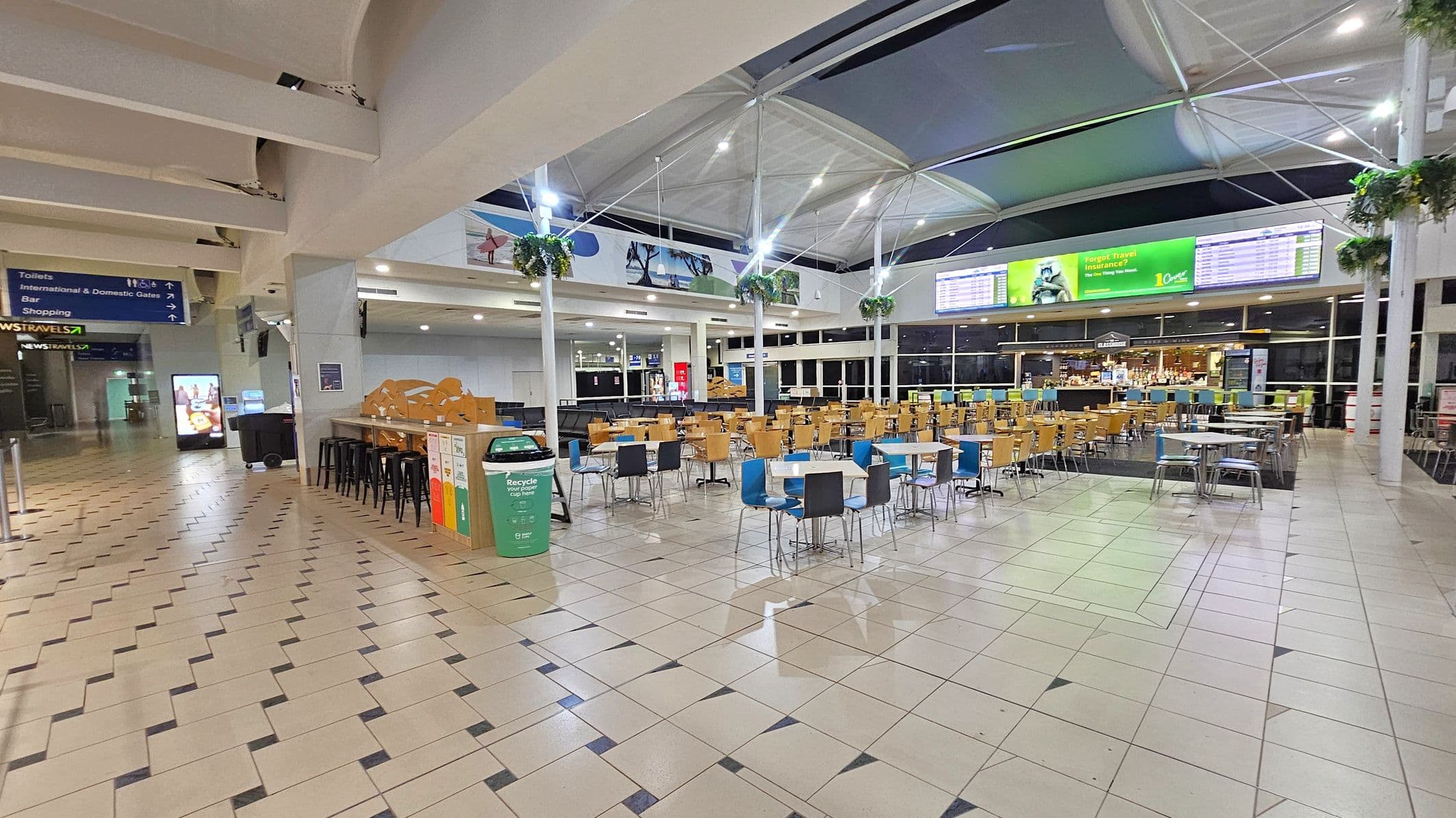 A spacious airport terminal with a tiled floor and high, white ceiling. On the left, there are signs directing to toilets, gates, a bar, and shopping. A vending machine and a newsstand are visible. In the centre, there are rows of tables and chairs, some with blue and black seats. Recycling bins are nearby. The right side features a large digital display board and a café counter. Hanging plants are suspended from the ceiling.
