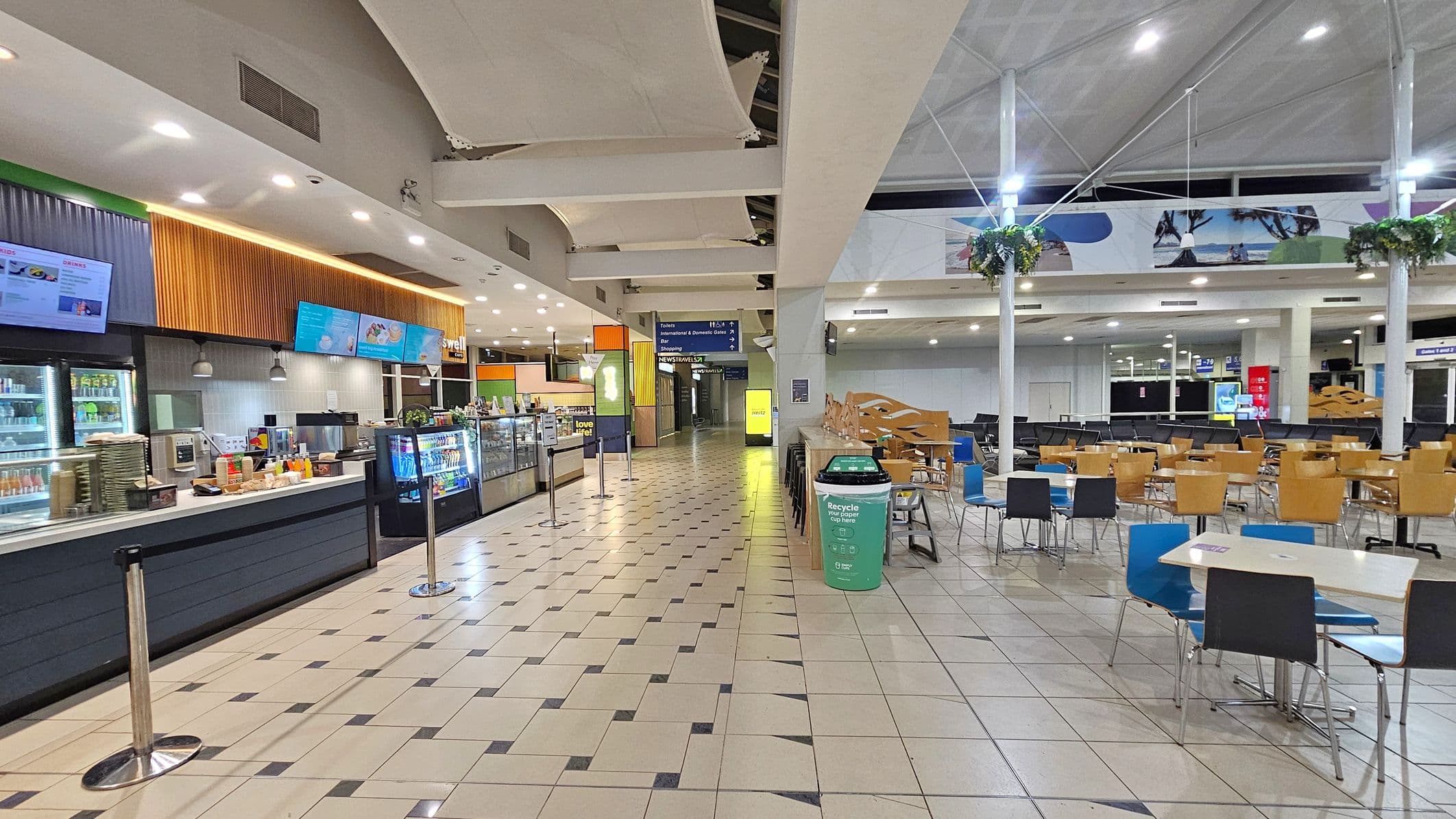 A spacious indoor area with a food court on the left featuring a long counter with display screens and a variety of food and drink items. The floor is tiled with a geometric pattern. On the right, there are tables and chairs in various colours, with a green recycling bin nearby. The ceiling is high with exposed beams and hanging lights. In the background, there are signs and more seating areas, with some decorative plants hanging from the ceiling.