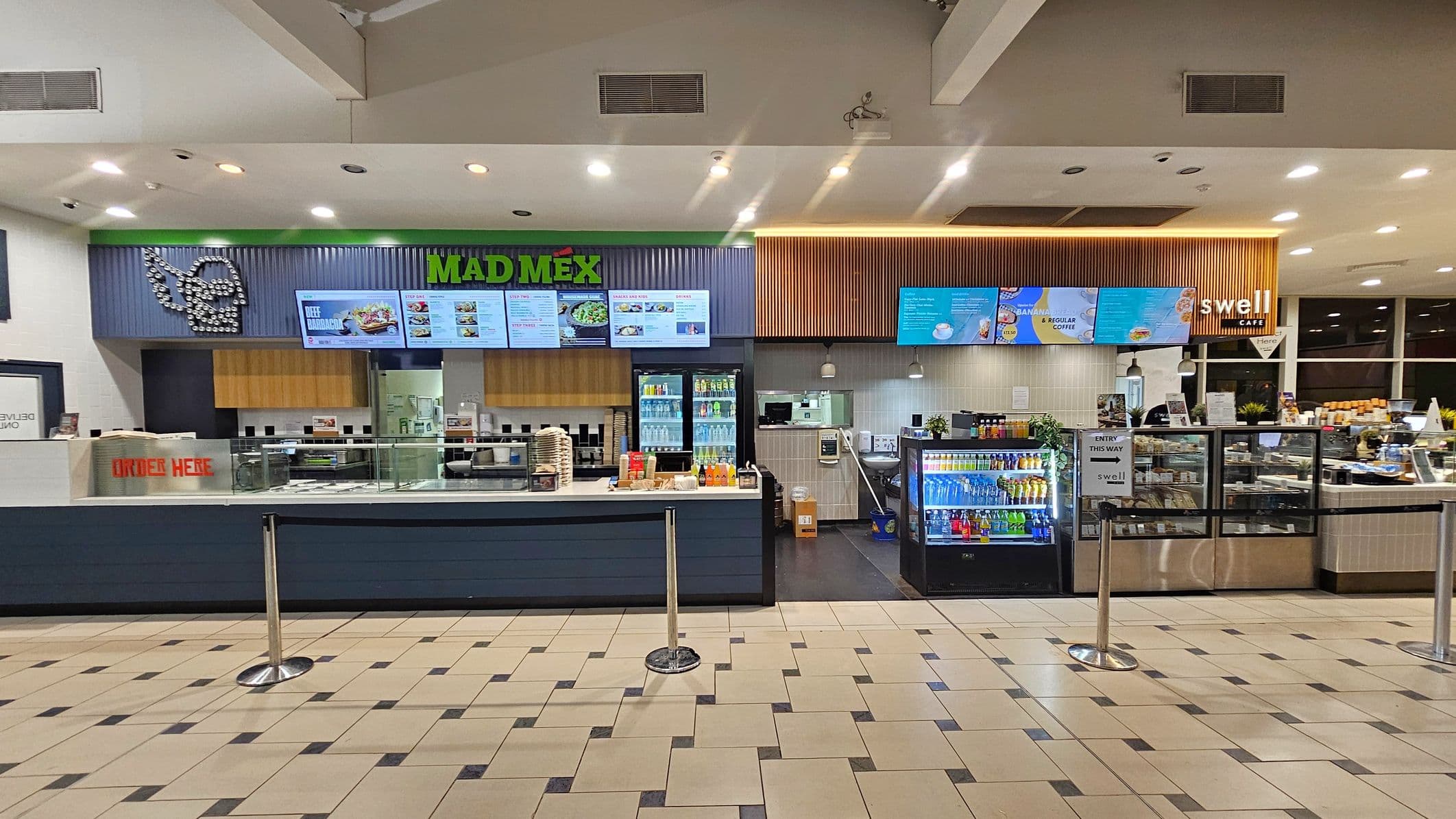 A food court with two distinct sections. On the left, "Mad Mex" features a corrugated metal facade with a logo and digital menu screens displaying food options. Below, a counter with a "Order Here" sign and a drinks fridge. On the right, "Swell Café" has wooden panelling, digital screens, and a counter with a variety of drinks and snacks. The floor is tiled in a beige and black pattern, and the ceiling has recessed lighting.
