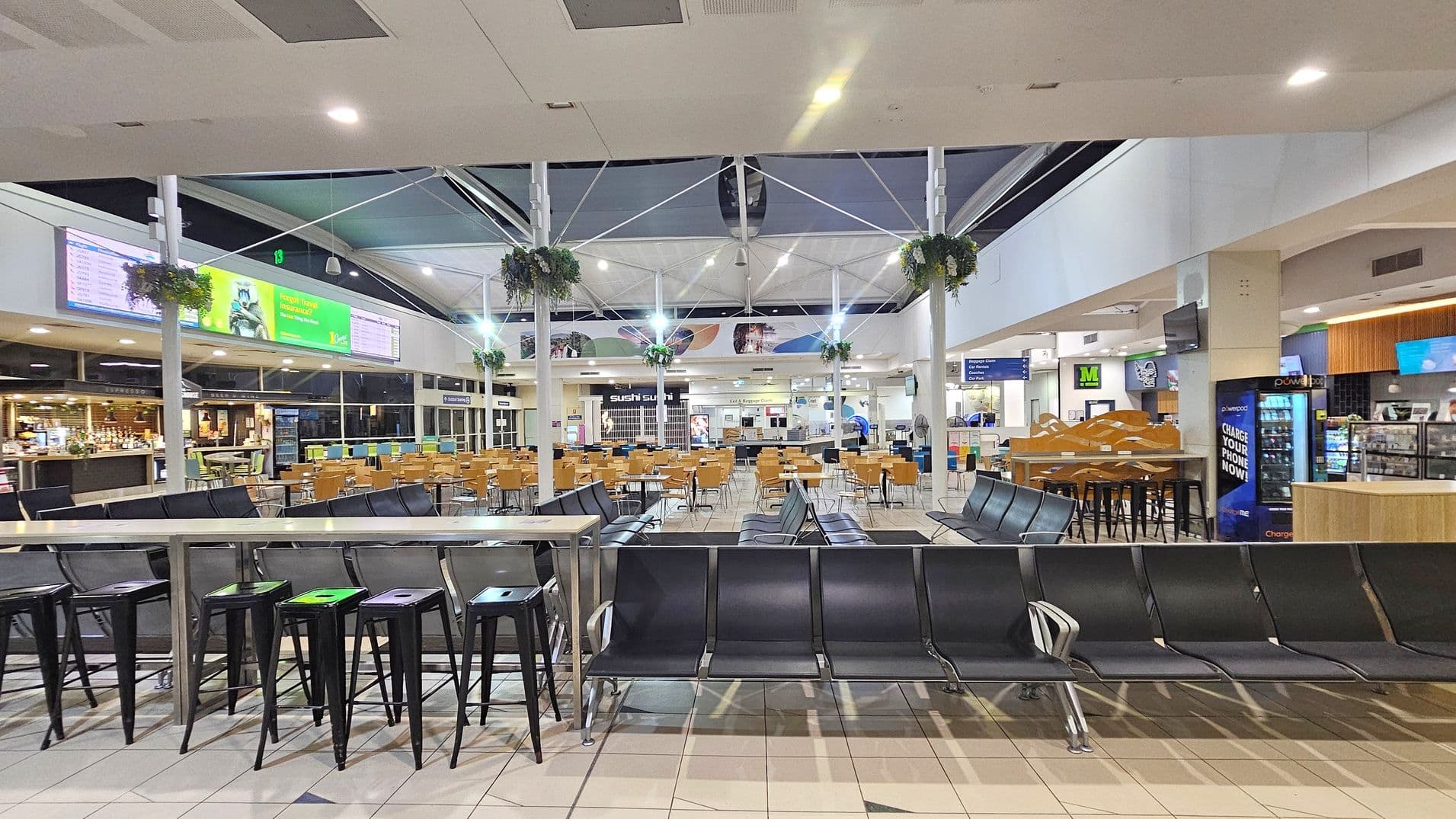 A spacious indoor area with a high ceiling featuring a network of white beams and hanging plants. In the foreground, there are rows of black seating and high stools. The middle section has numerous wooden tables and chairs. To the left, there's a bar with a counter and stools. On the right, a convenience store with a sign for phone charging. Digital screens and advertisements are visible on the walls. The floor is tiled in a light colour.