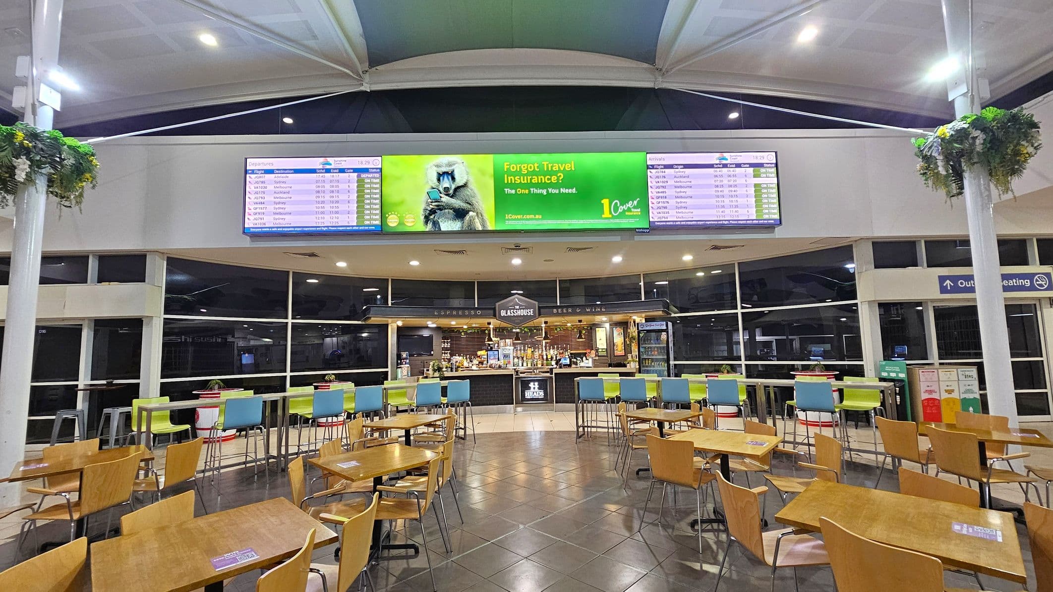 Inside an airport terminal, there are wooden tables and chairs arranged in the foreground. In the middle, a bar area named "Glasshouse" offers espresso, beer, and wine, with colourful high stools in front. Above, digital screens display flight information and an advertisement about travel insurance. The ceiling is high with a modern design, and hanging plants are attached to white pillars on both sides. The floor is tiled, and large windows are visible in the background.