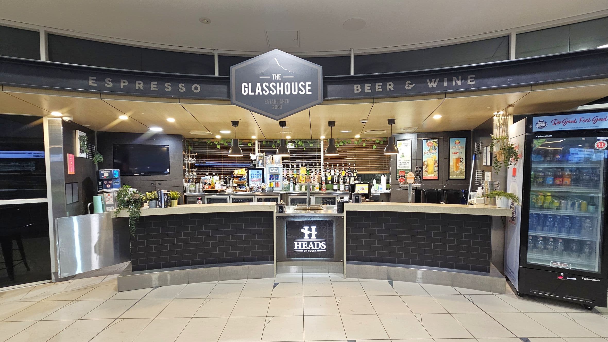 A bar area with a sign reading "The Glasshouse" above. The counter is curved with a black tile front and a "Heads" logo. Behind the counter, there are various bottles and a coffee machine. To the right, a glass-fronted refrigerator displays drinks. The ceiling has recessed lighting and hanging lamps. The floor is tiled in a light colour. Potted plants are placed on the counter and hanging on the right.