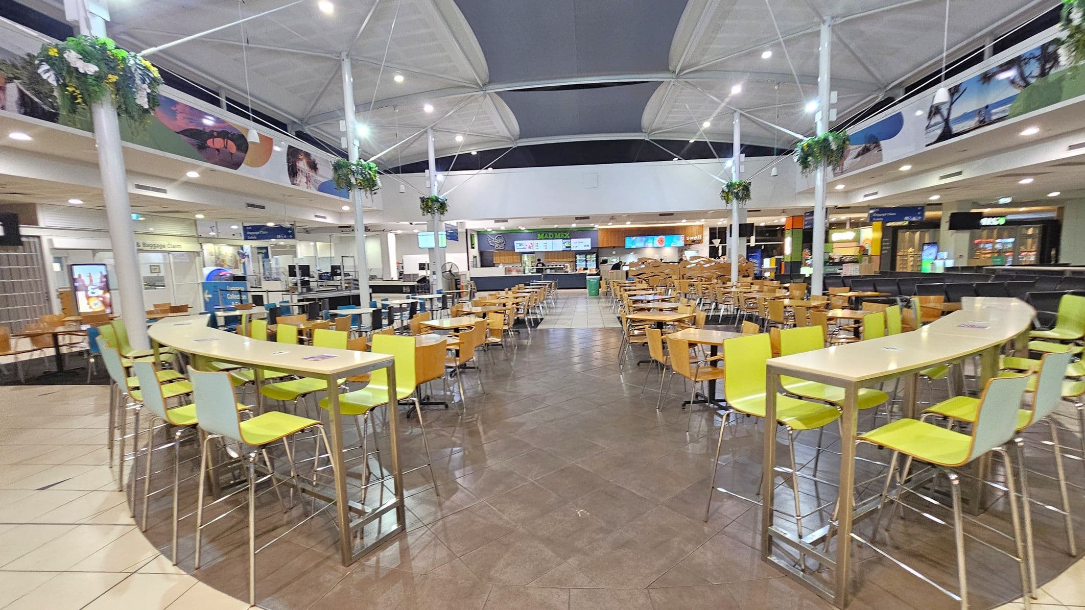A spacious food court with a high, white ceiling featuring geometric patterns. In the foreground, there are curved tables with green and light blue chairs. The middle area has numerous wooden tables and chairs arranged neatly. The background shows various food outlets with illuminated signs. Hanging plants are suspended from the ceiling, and the floor is a mix of tiled patterns. Bright lighting illuminates the entire area.